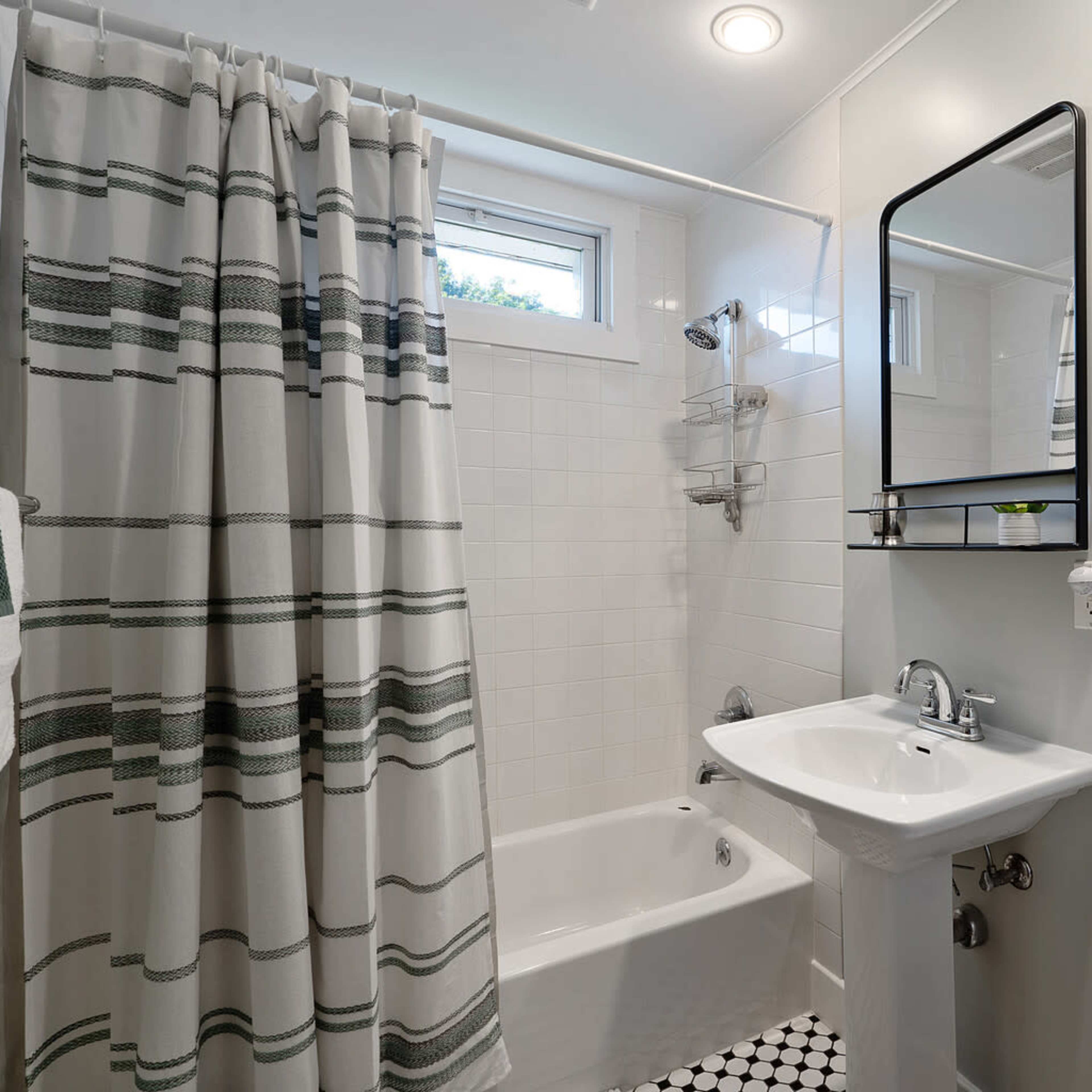 A small, modern bathroom featuring a bathtub with a shower, a pedestal sink, a mirror, and a white tile design with black accents.