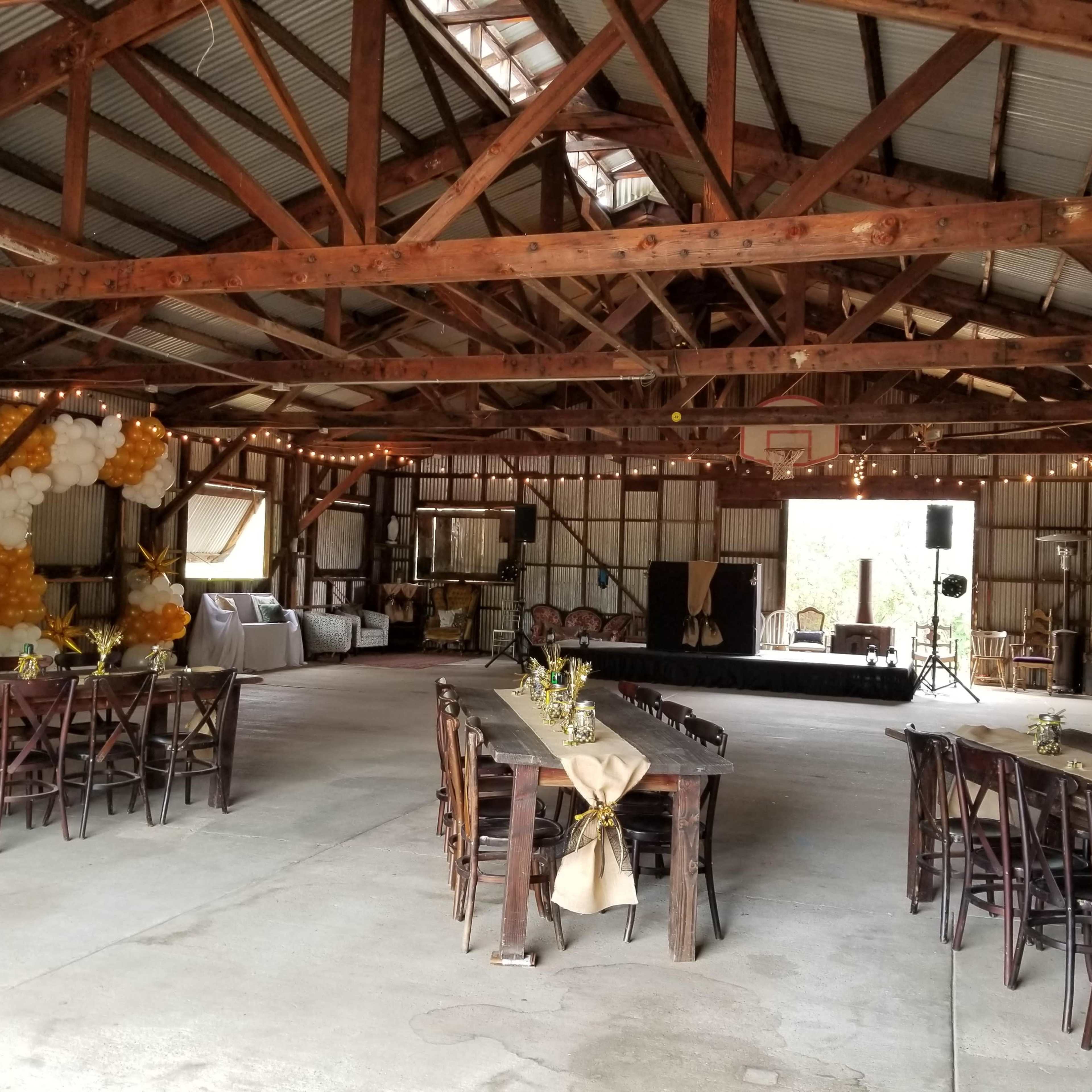 The interior of a rustic barn is decorated for an event, featuring wooden tables, string lights, and a stage area.