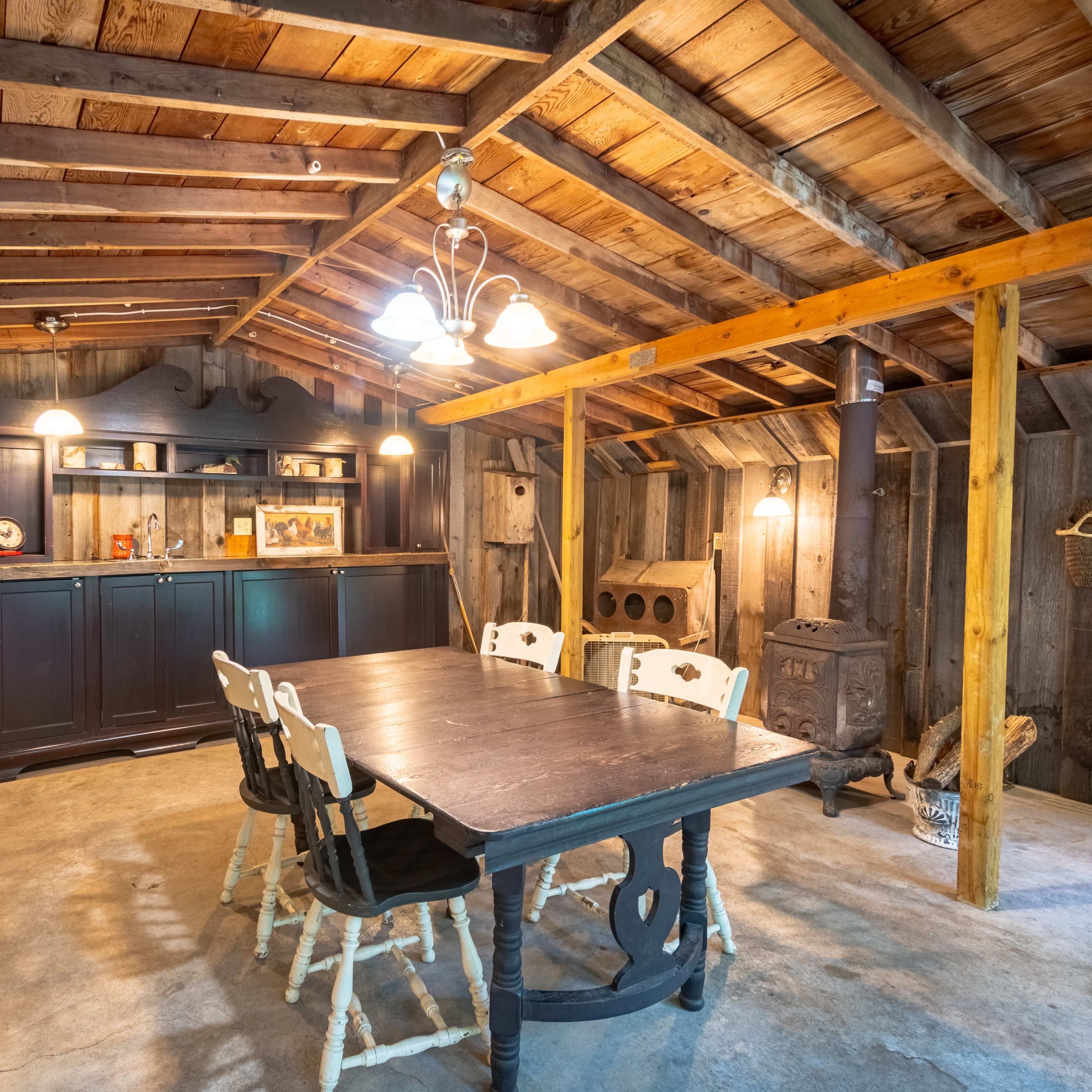 The interior of a rustic cabin features a wooden table with white chairs, a dark wooden bar unit, and a stove against the wall beneath a wooden ceiling.