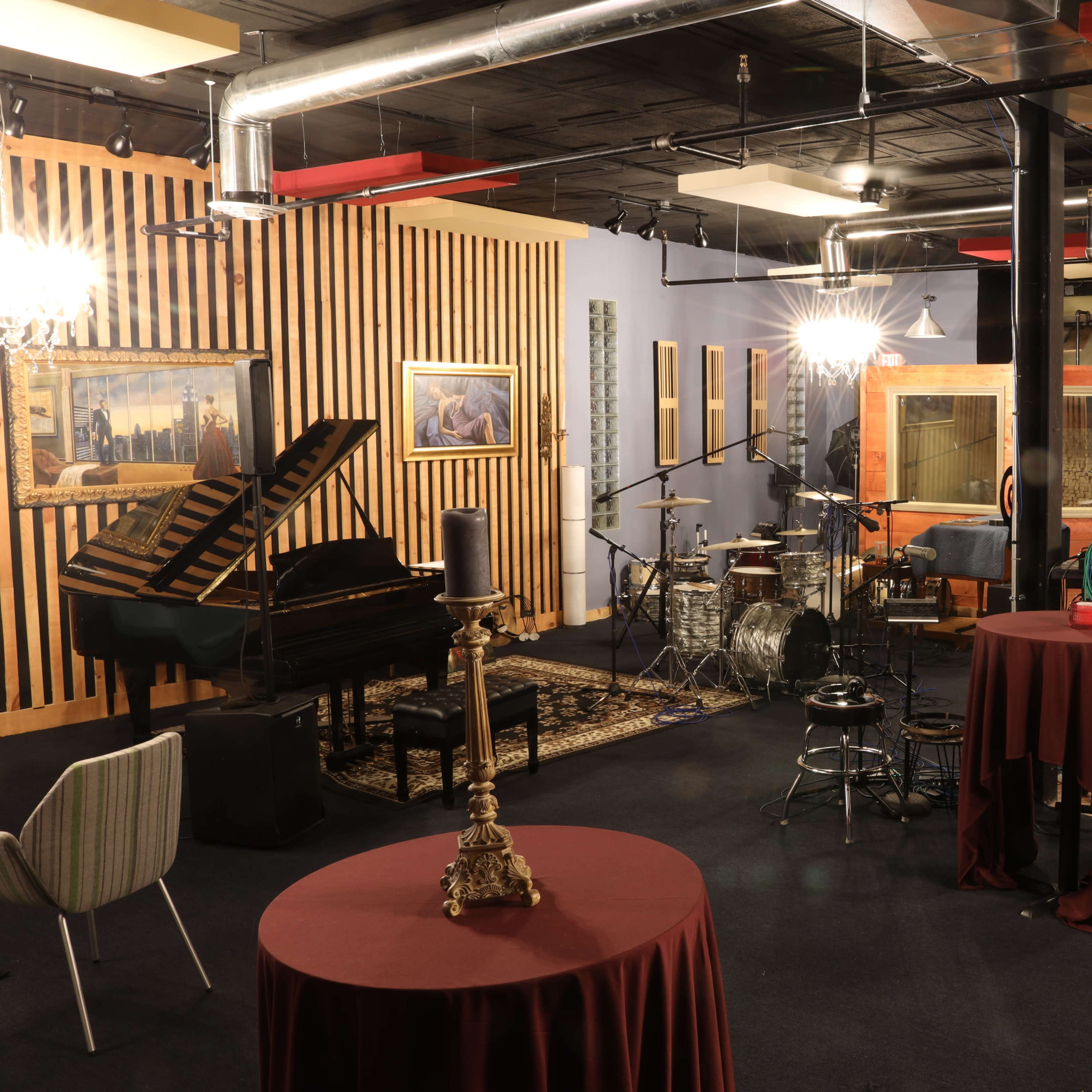 A music studio featuring a grand piano, a drum set, and several tables, with a cozy seating area and kitchen in the background.