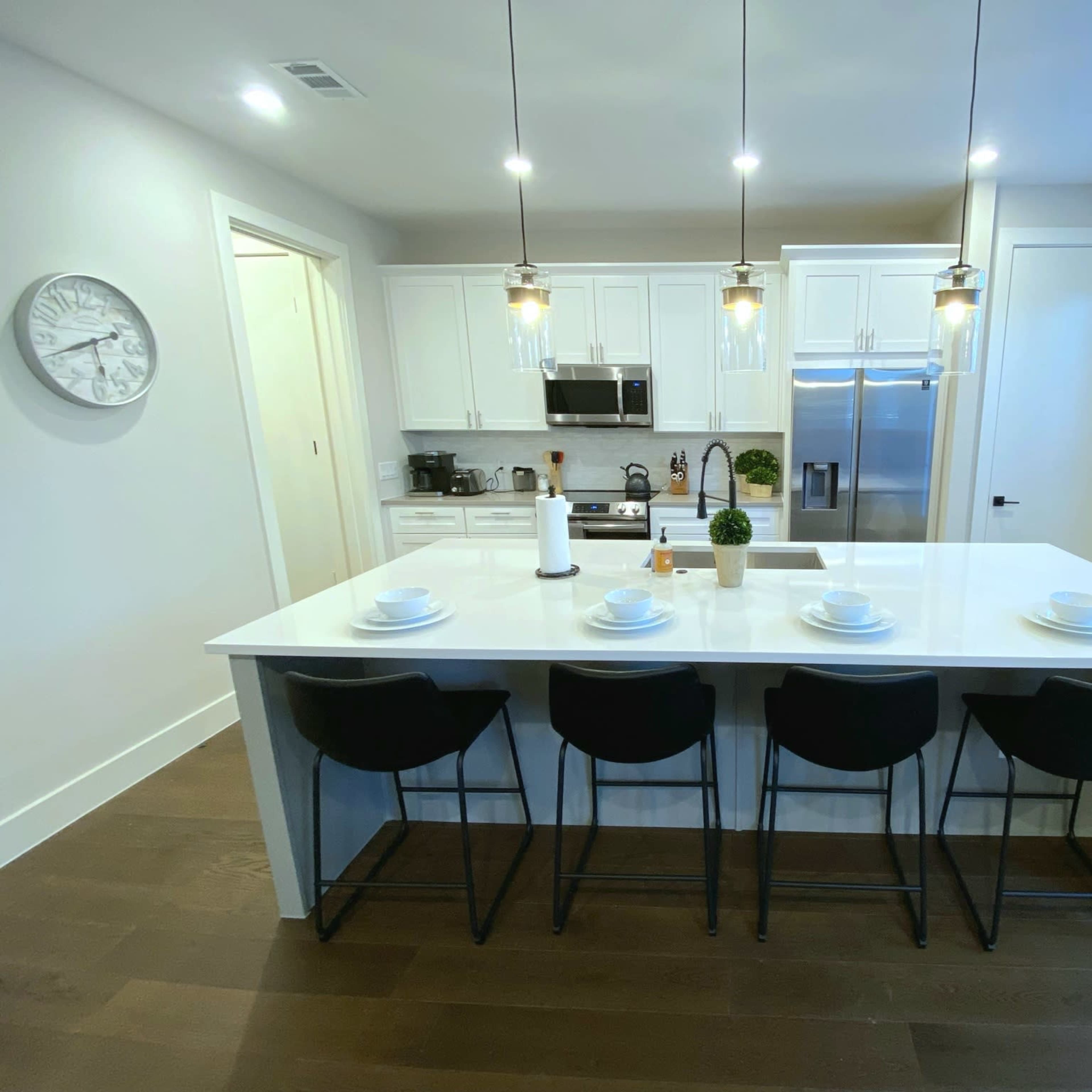 A modern kitchen features a large island with four black bar stools, white cabinetry, stainless steel appliances, and pendant lighting.