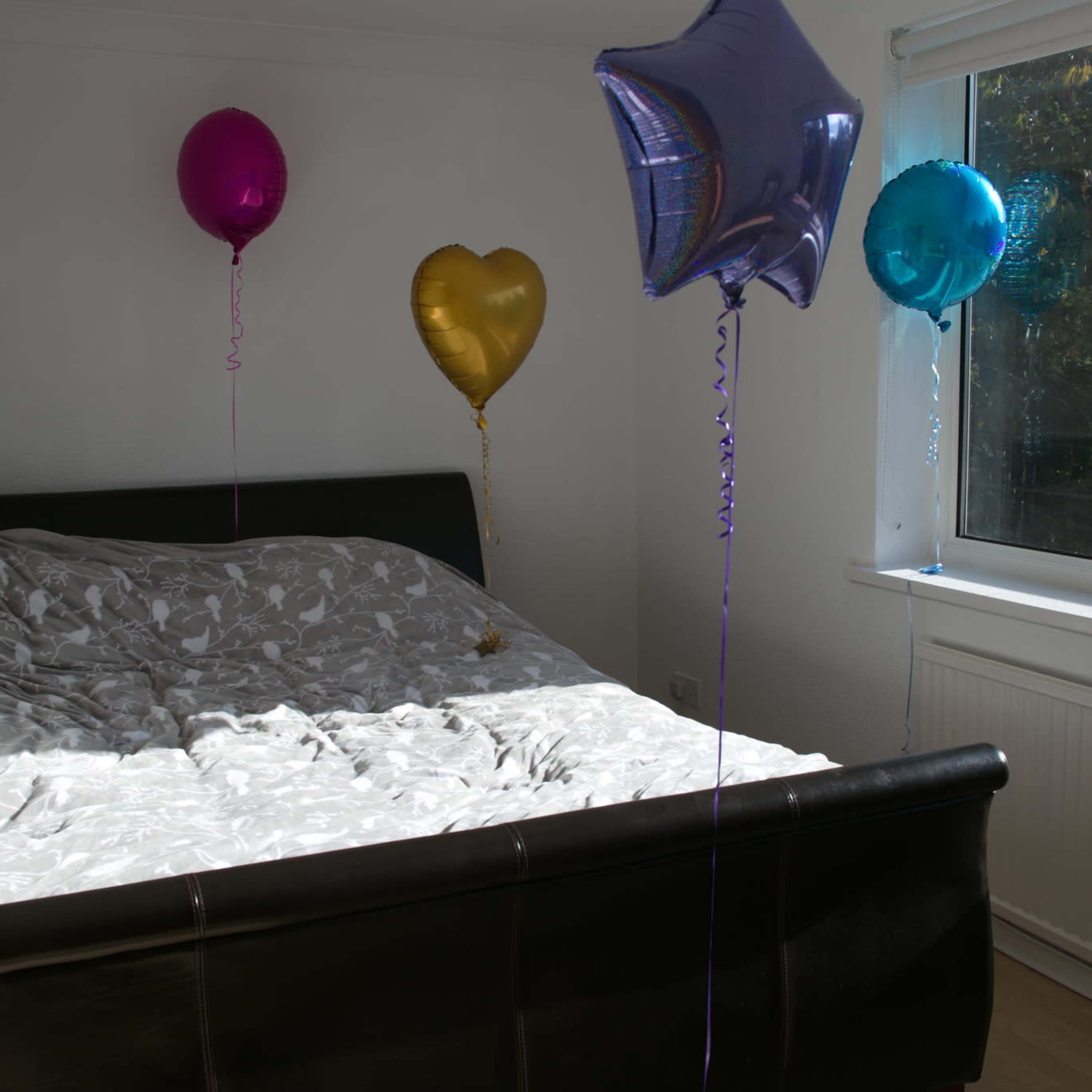 A bed with a patterned cover is positioned in a well-lit room, surrounded by colorful balloons of various shapes.