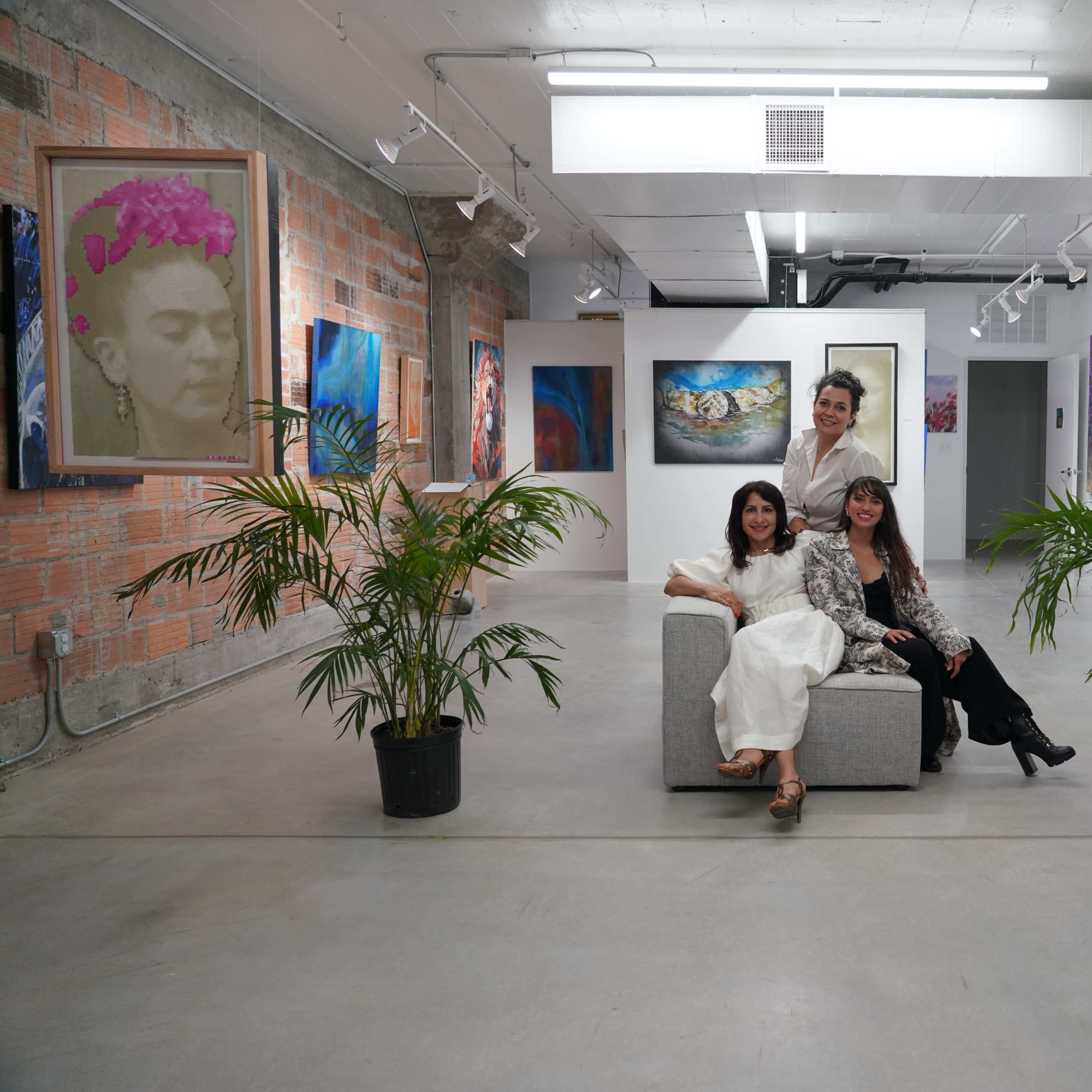 Three women pose on a gray sofa in an art gallery filled with colorful paintings.
