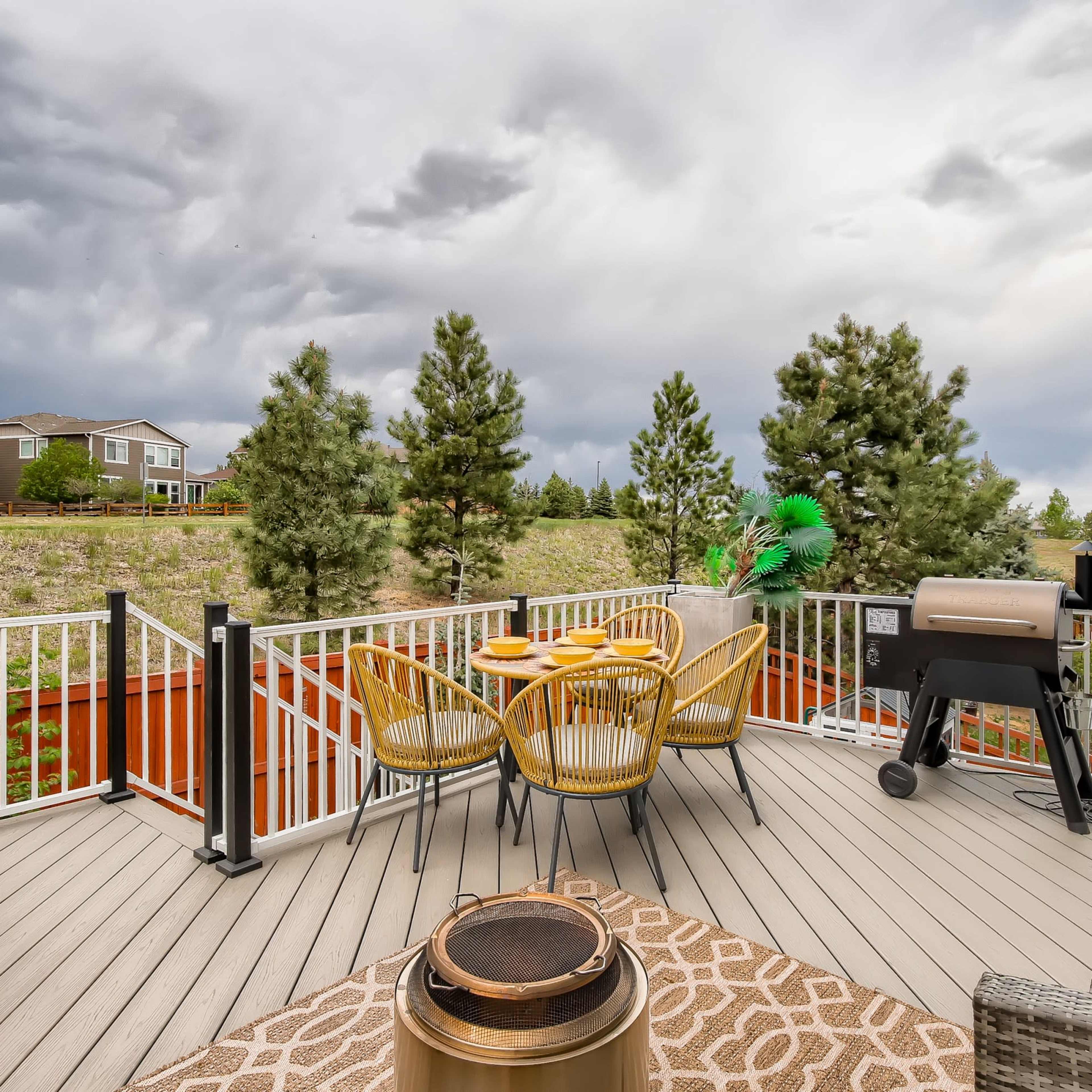 A deck features a table with chairs, a grill, and a fire pit, overlooking a grassy area with trees and houses in the background under a cloudy sky.