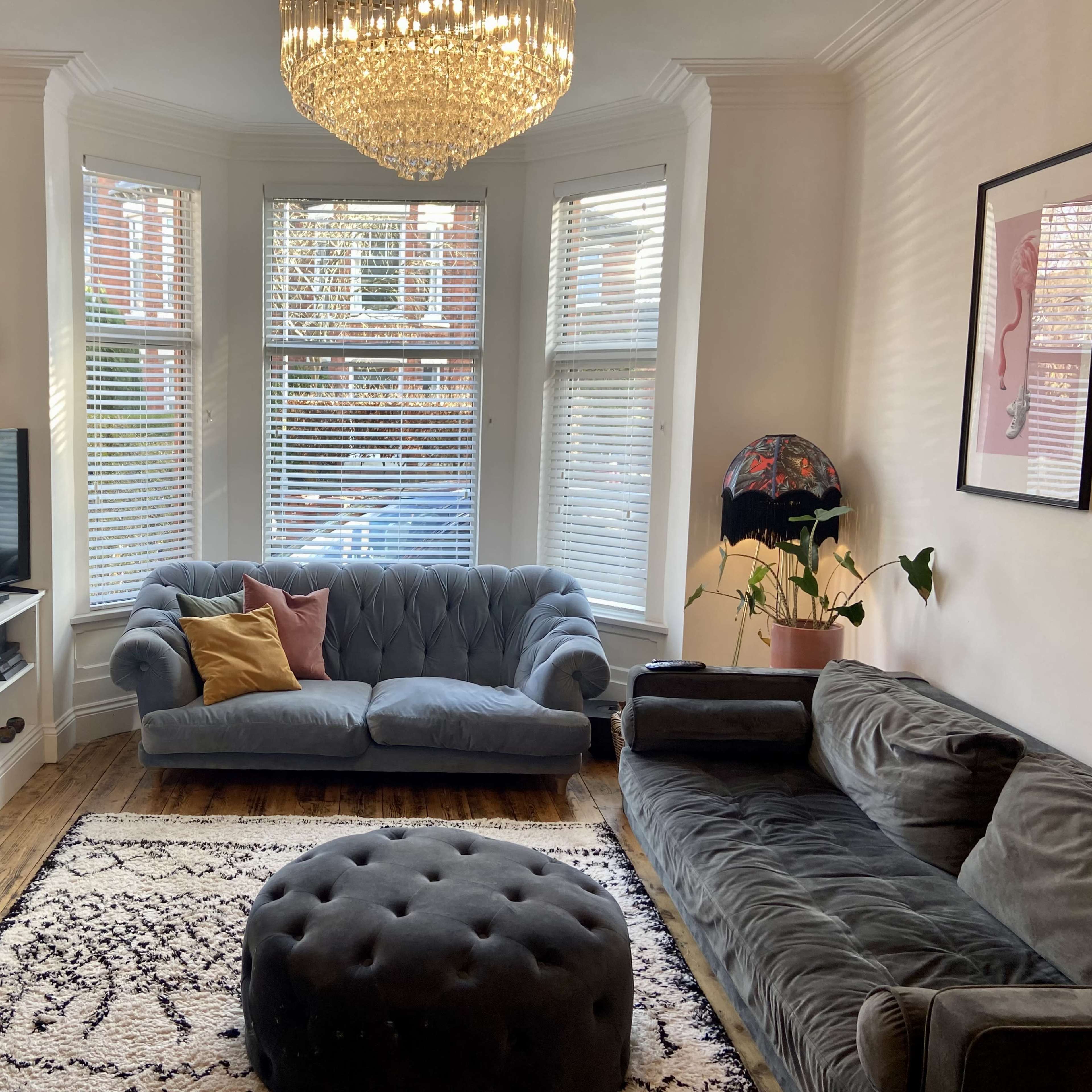 The room features a stylish seating area with a tufted blue sofa and a large round ottoman, illuminated by a chandelier and natural light from bay windows.
