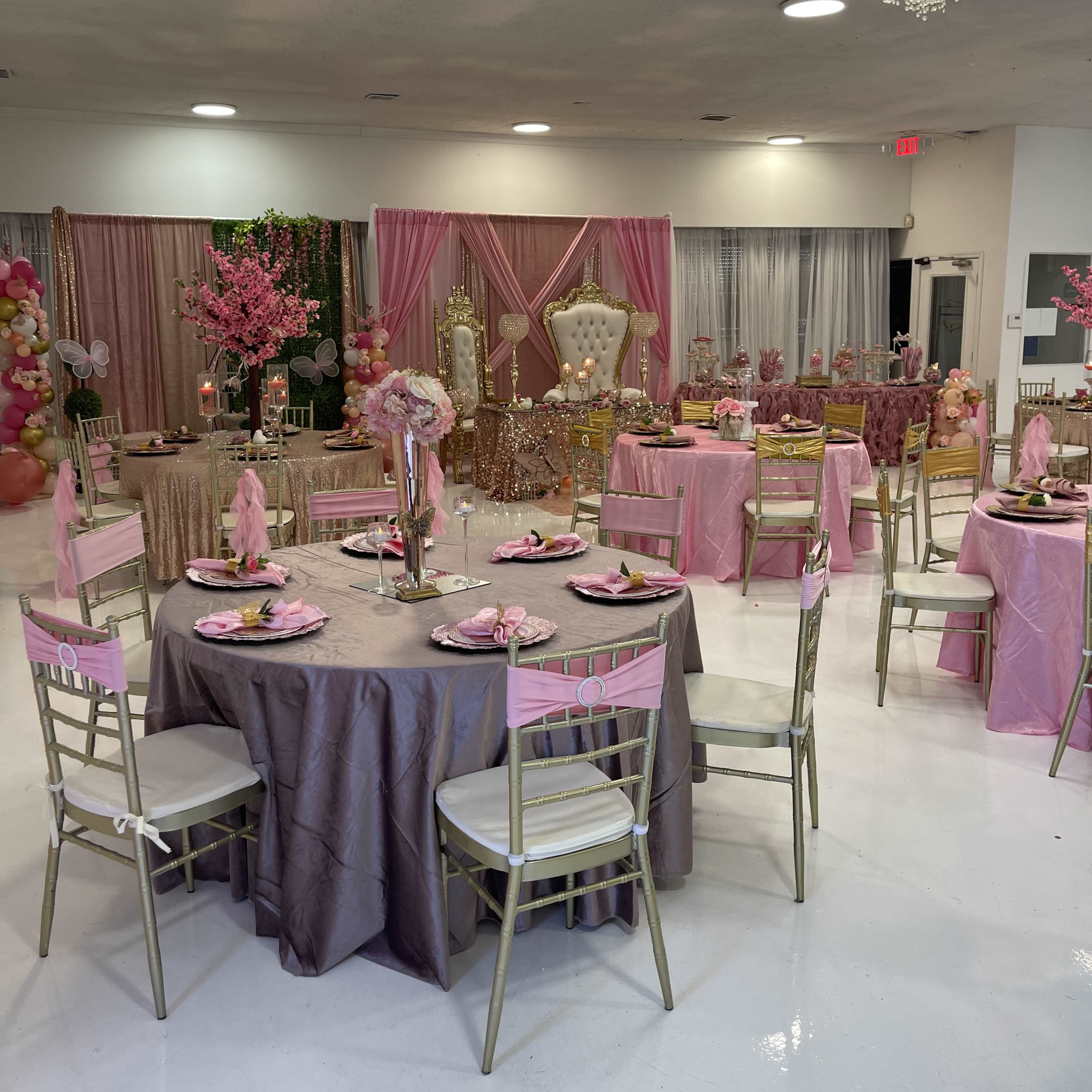 A decorated event space featuring tables set with pink and gold tableware, a large chandelier, and a backdrop of flowers and ornate furniture for a celebration.