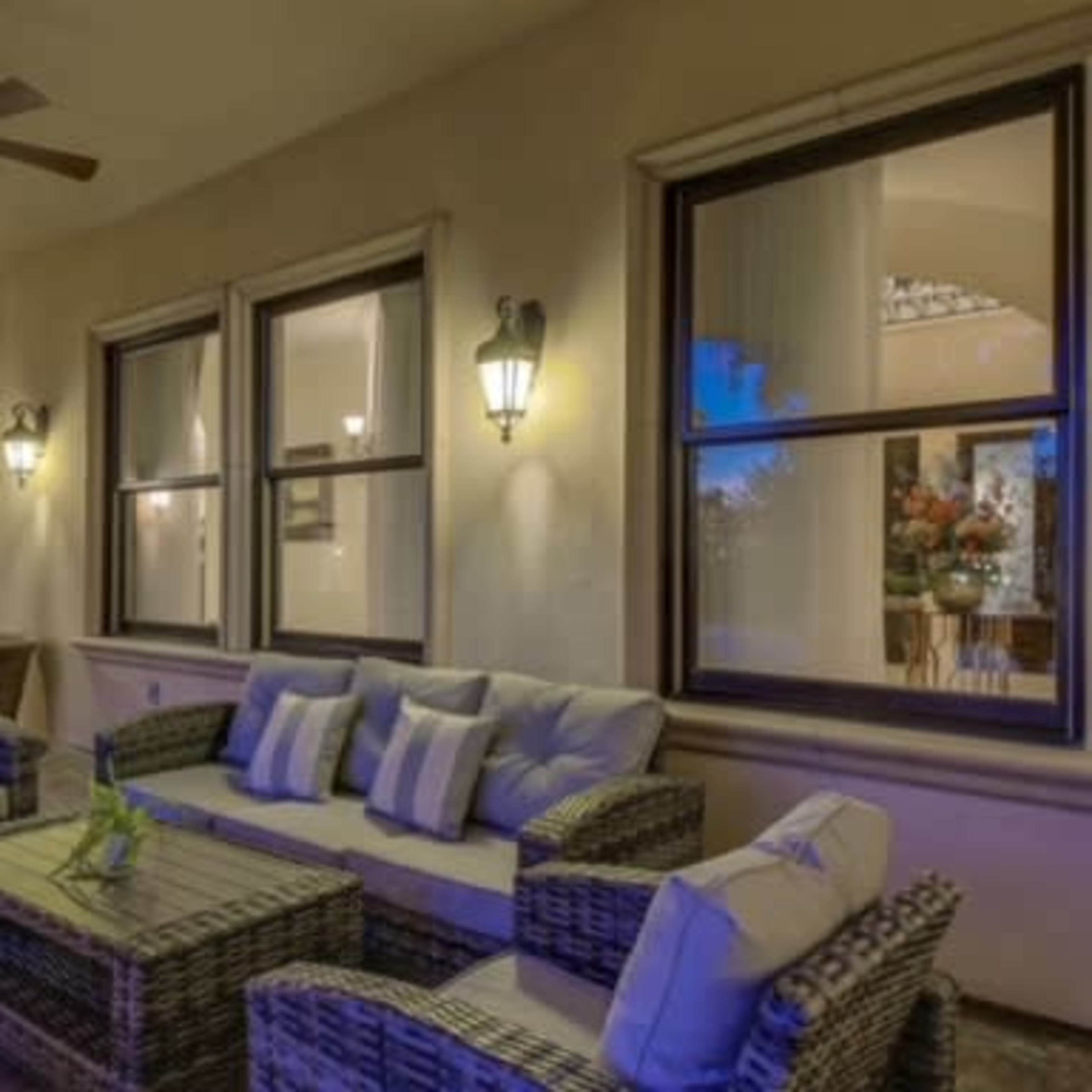 A covered outdoor seating area features a wicker couch and table, illuminated by wall sconces and large windows.