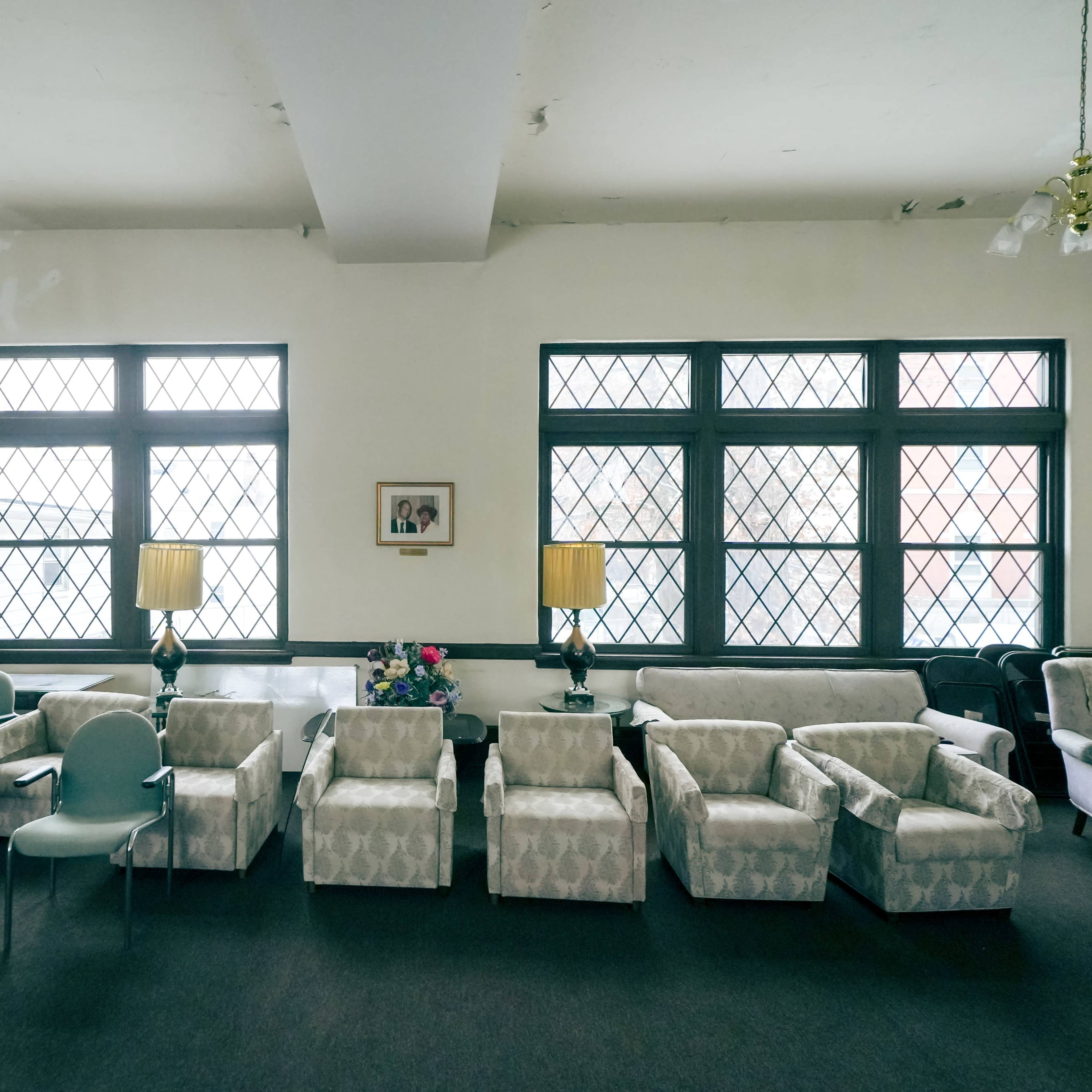A spacious room with several chairs and armchairs arranged in two rows facing each other, complemented by two lamps and a framed picture on the wall.