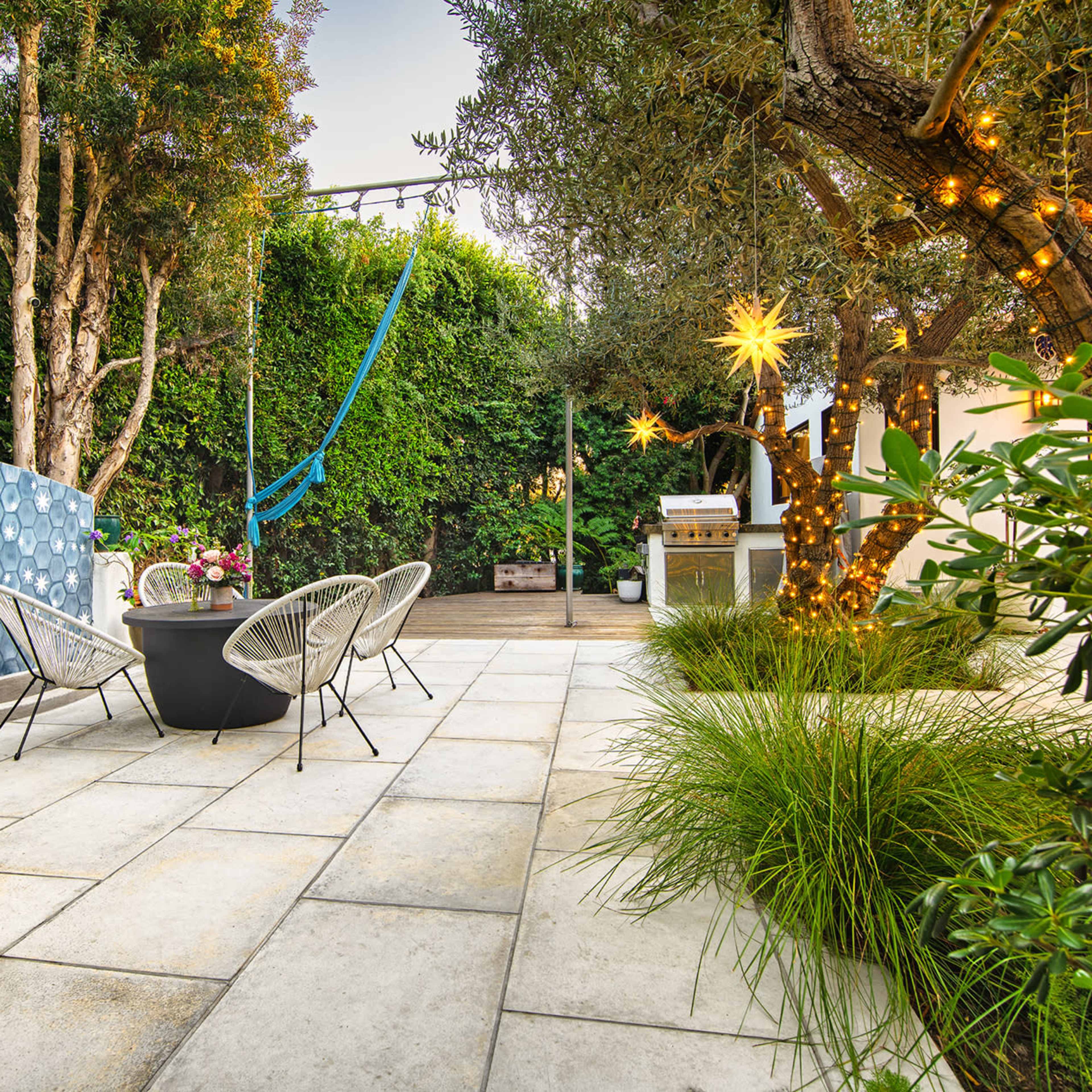 A landscaped outdoor patio area with stone tiles, potted plants, and a blue wall feature, accompanied by a seating arrangement of black chairs and a hammock nearby.