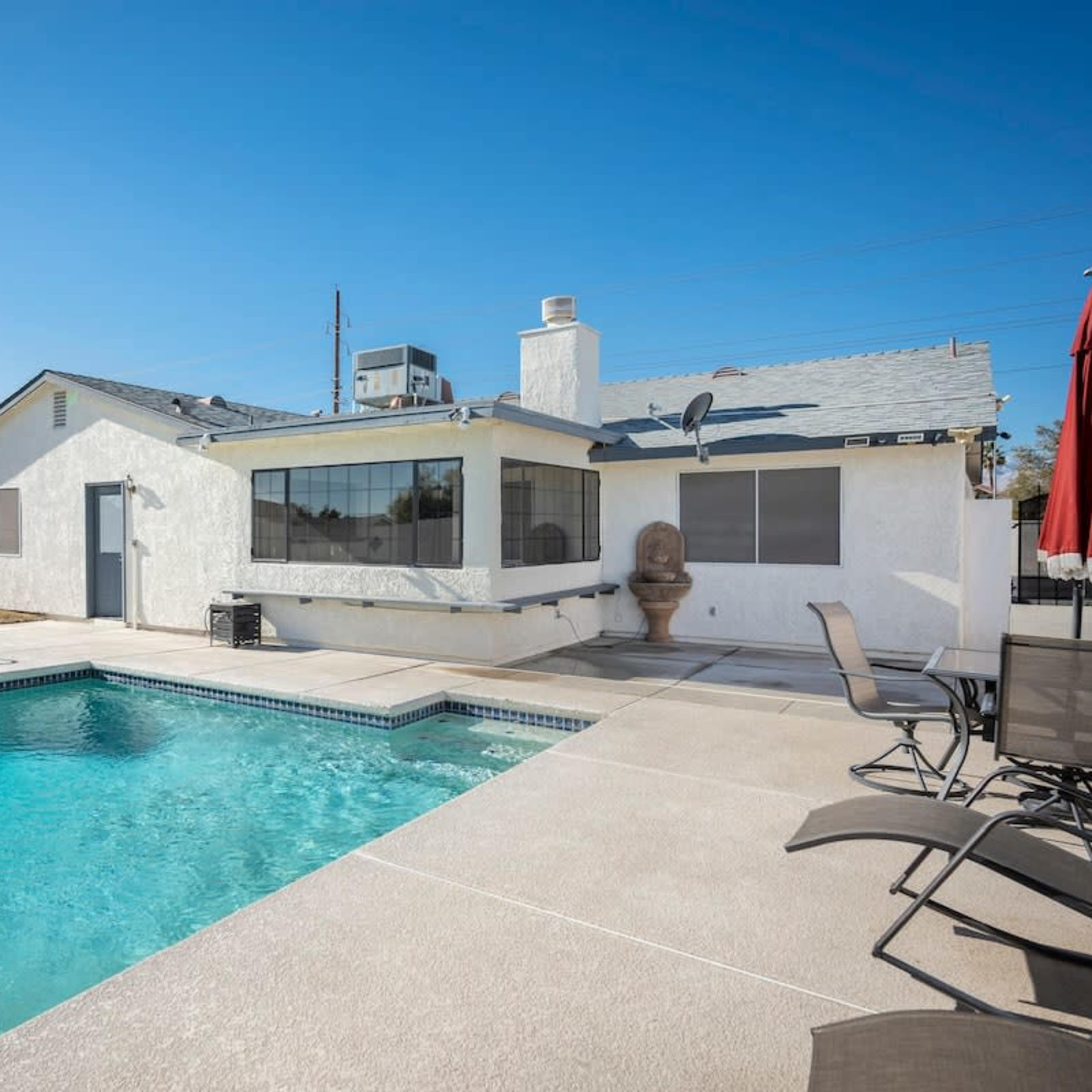 A backyard with a swimming pool adjacent to a single-story house featuring large windows and a concrete patio.