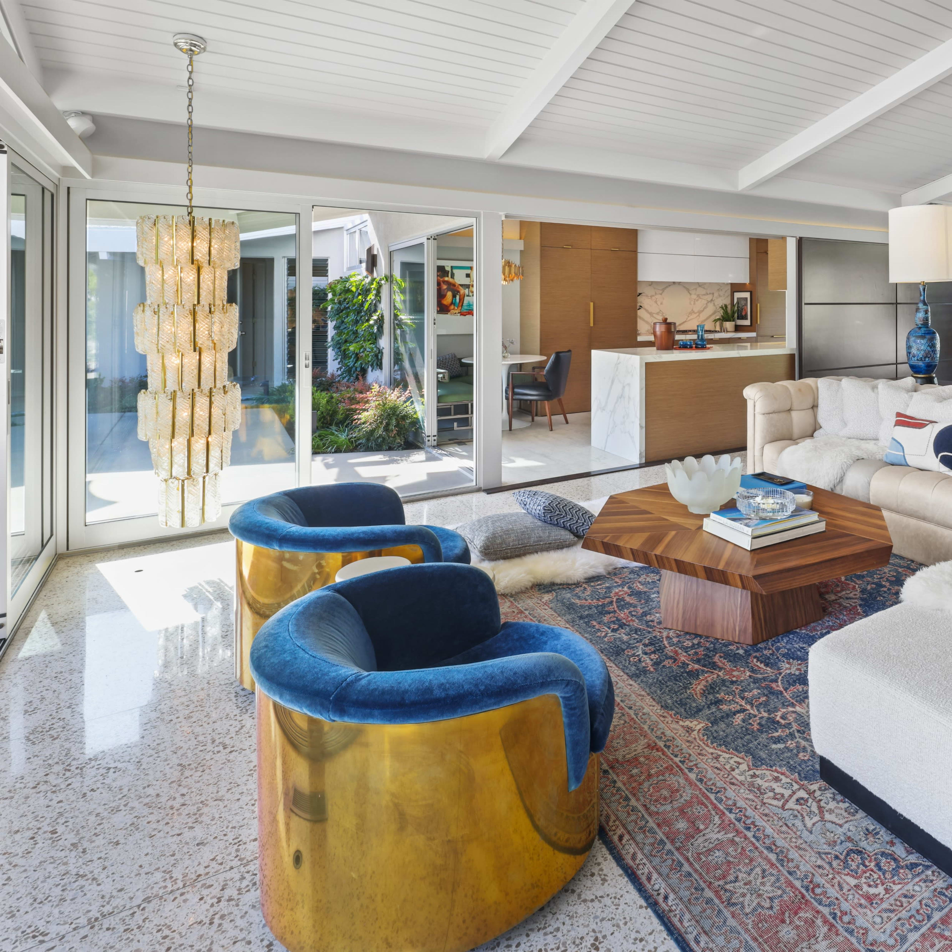 A modern living room with a light-colored sofa, two blue chairs, a wooden coffee table, and large windows that provide natural light.