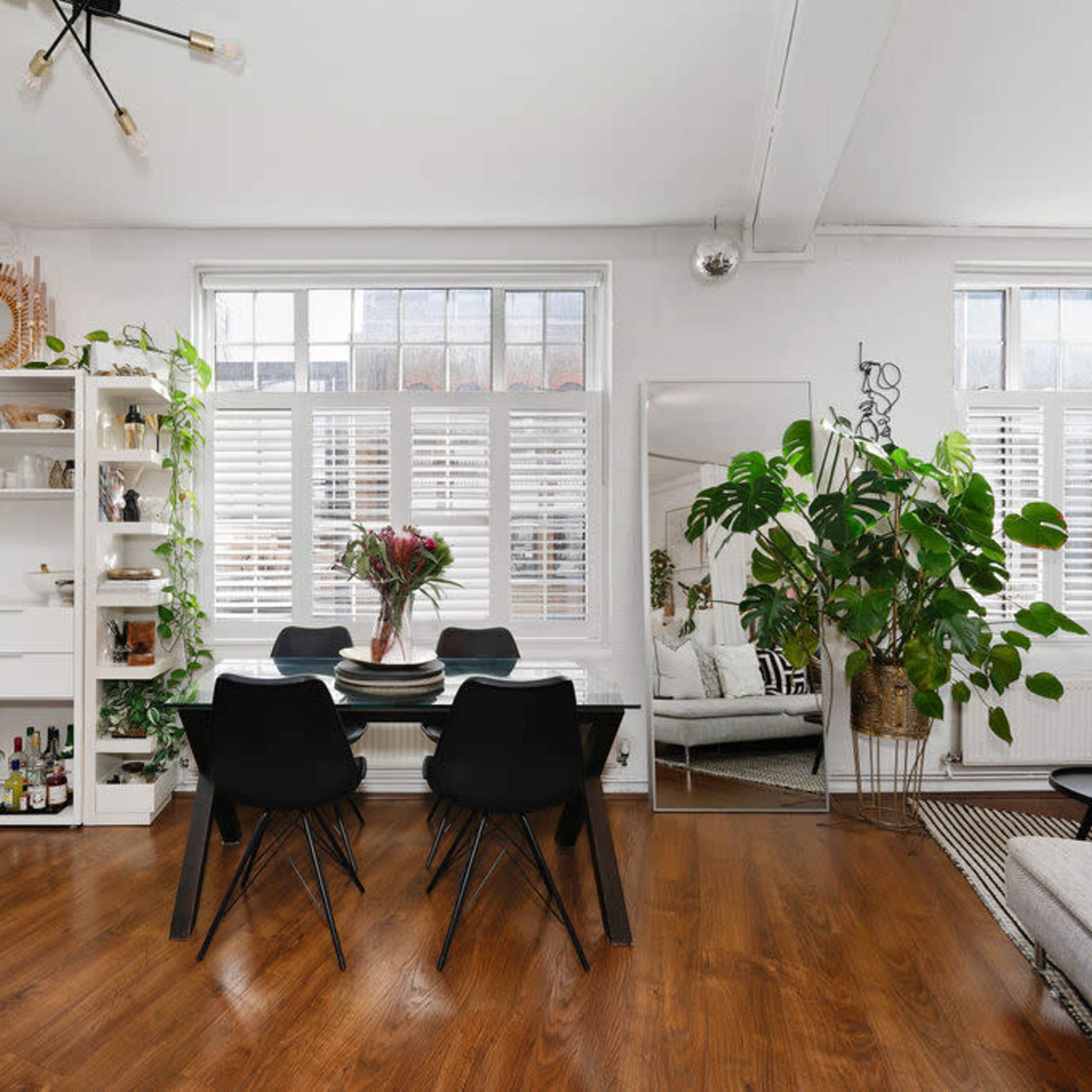 A modern dining area with a black table and chairs, surrounded by plants and shelves against a backdrop of large windows with wooden shutters.