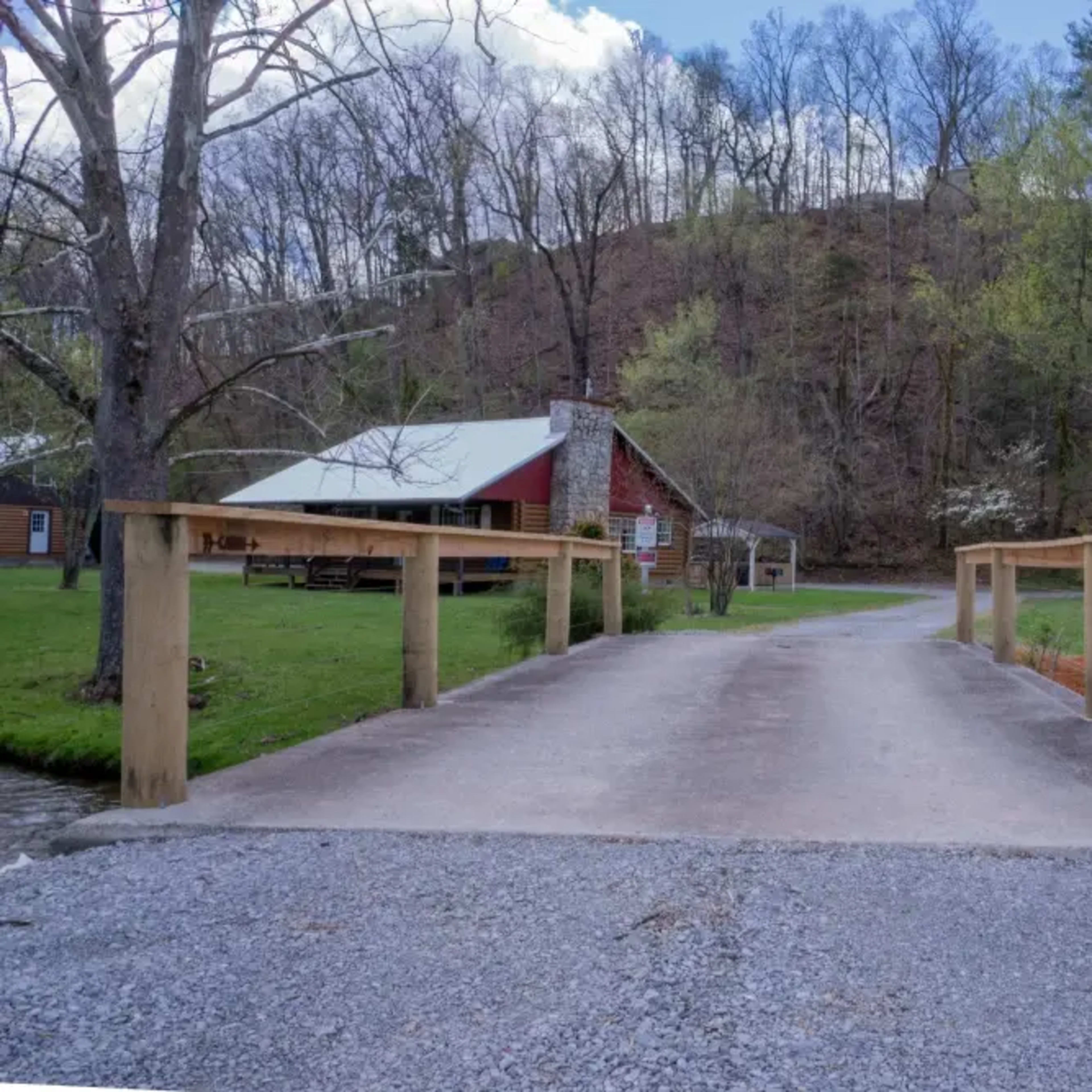 A gravel road leads to a wooden bridge crossing a small stream, with a cabin and trees visible in the background.