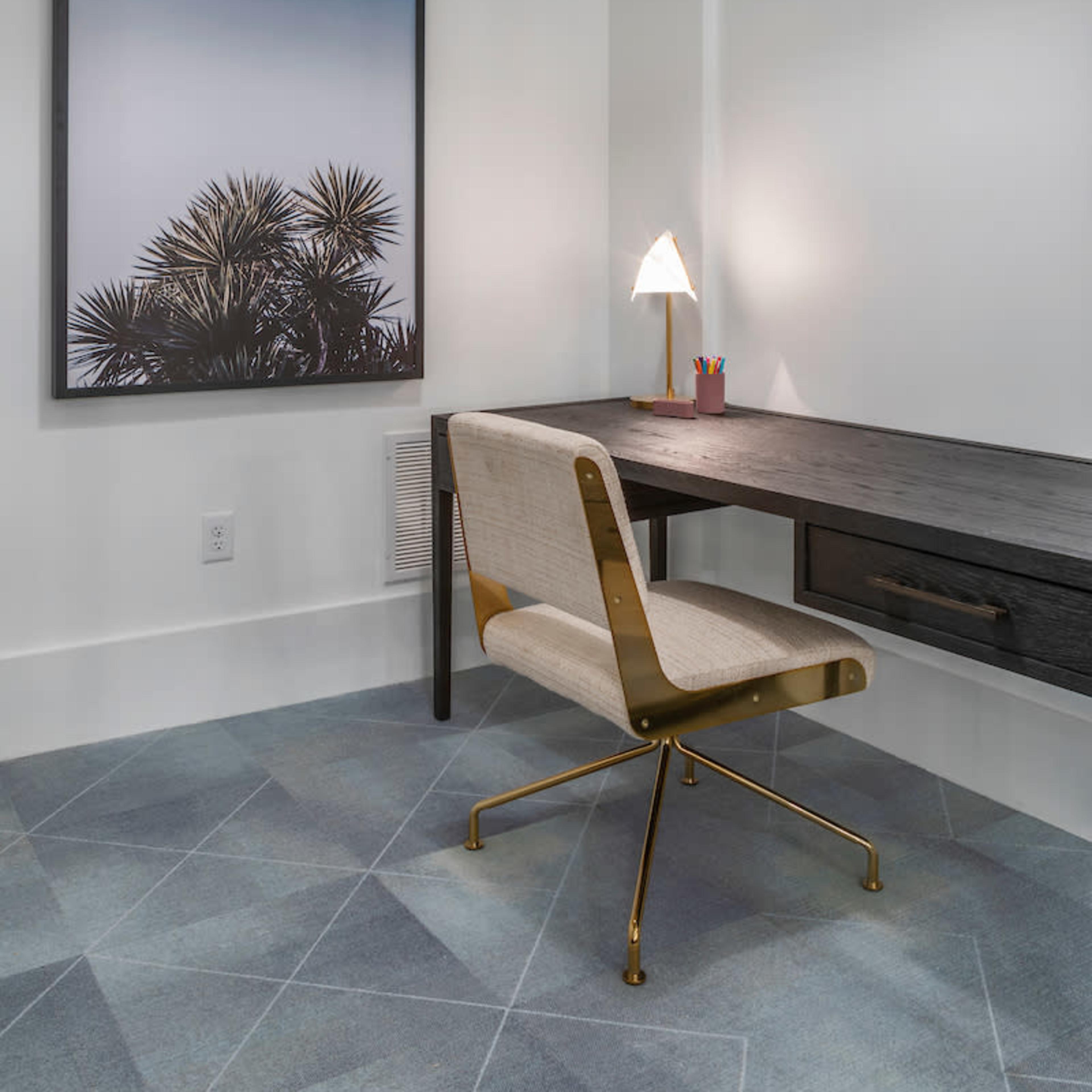 A modern workspace featuring a wall-mounted framed photograph, a simple desk with a drawer, a stylish chair, and a warm light fixture overhead.