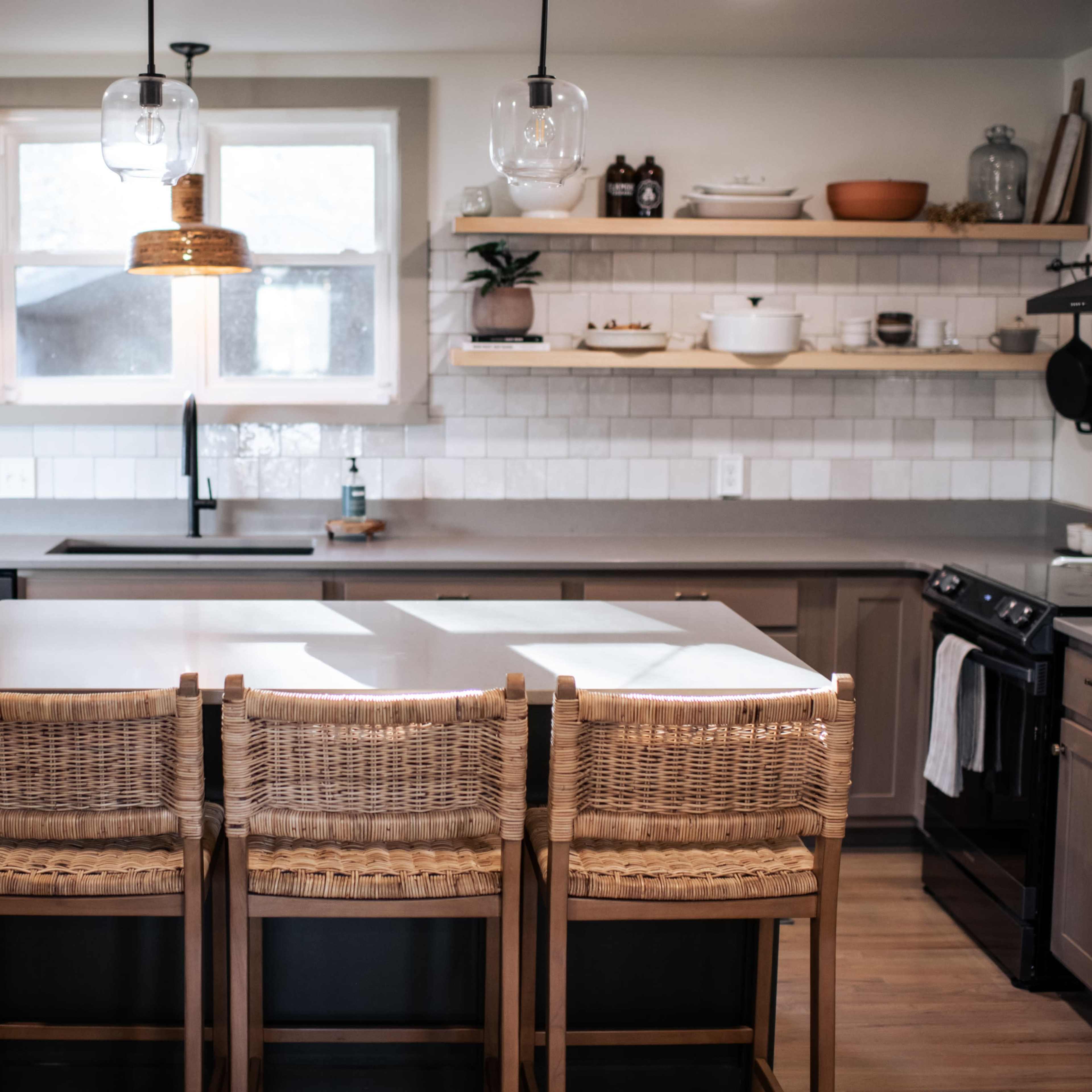 A modern kitchen features a large island with three woven bar stools, open shelving displaying kitchenware, and a window above the sink.