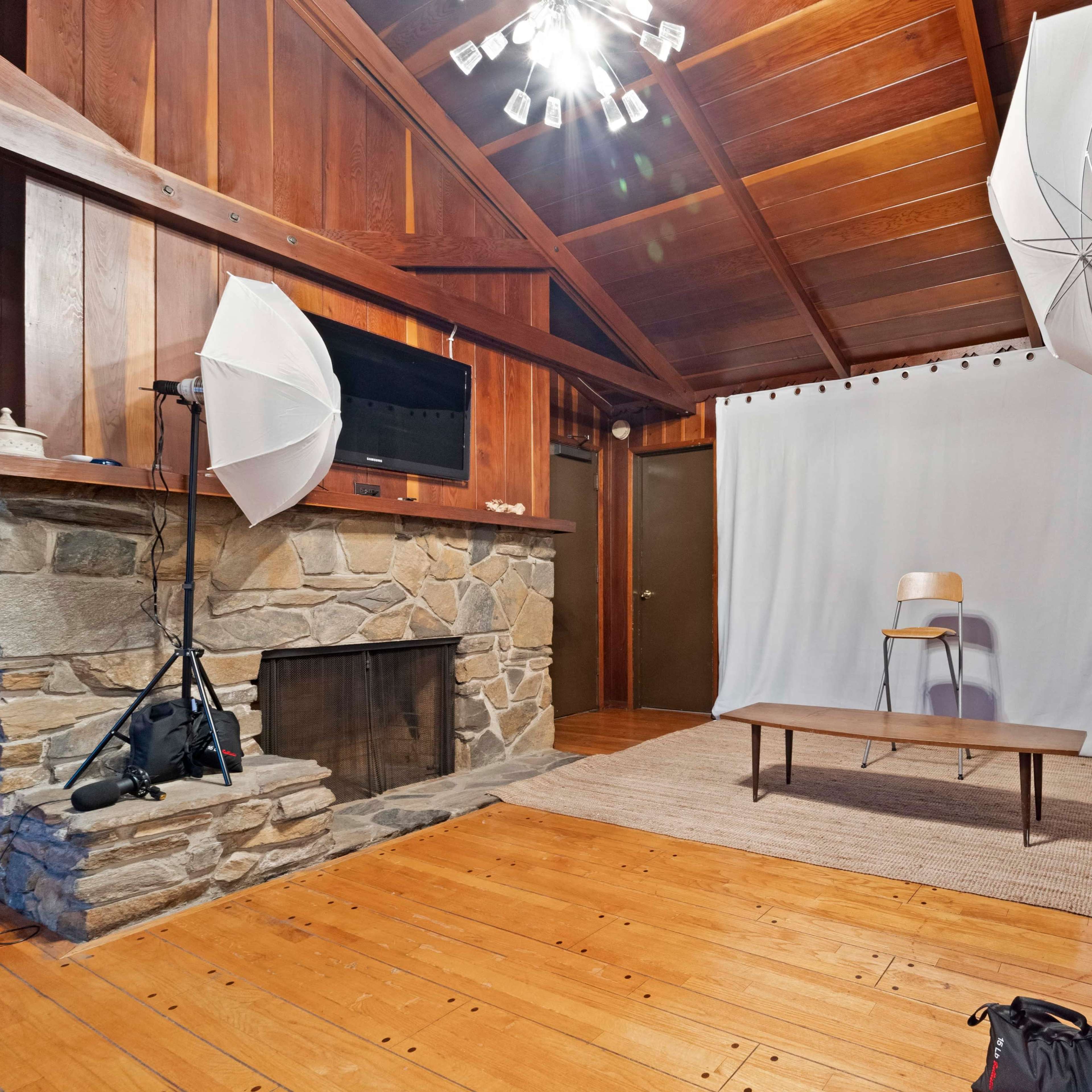 A room with wooden walls, a stone fireplace, a plain white backdrop with a wooden bench, and two softbox lights positioned on either side.