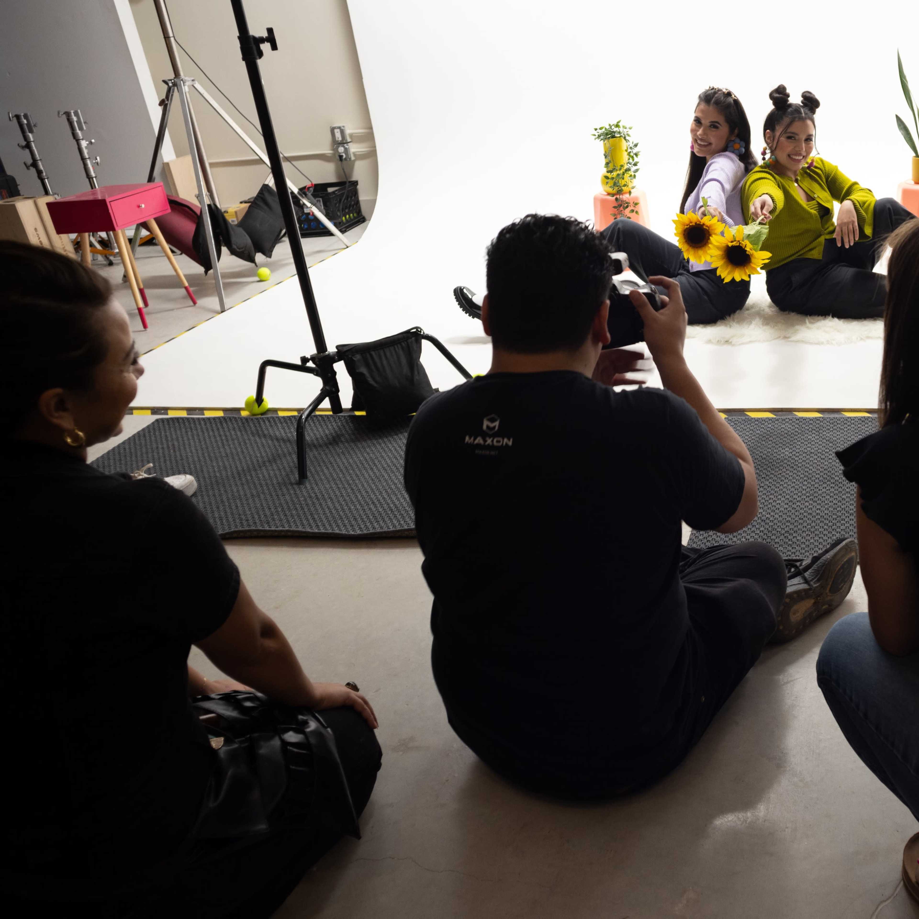 A photographer captures two models posing with sunflowers and plants in a well-lit studio.