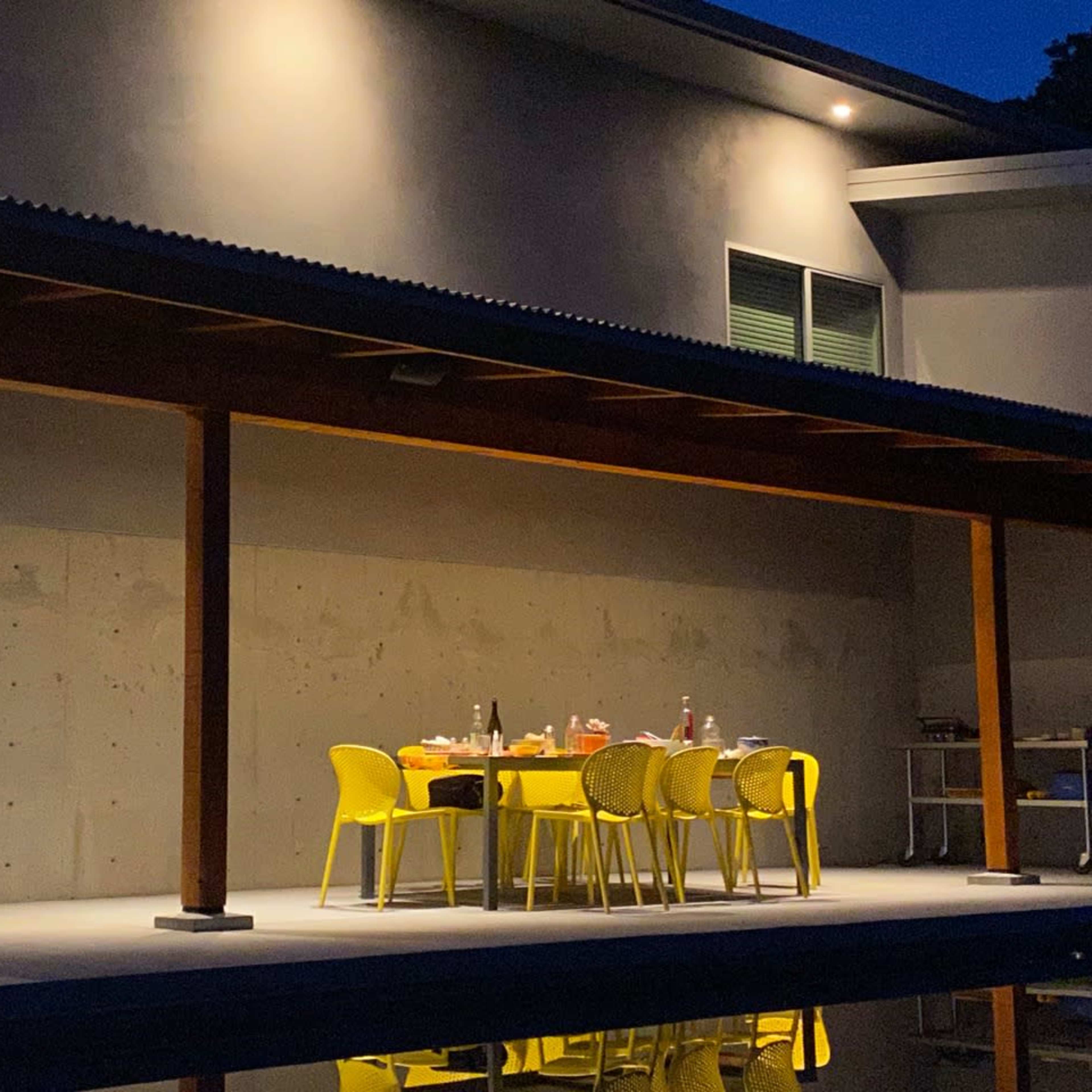 A table with yellow chairs is set under a covered patio next to a pool, illuminated by overhead lights at night.