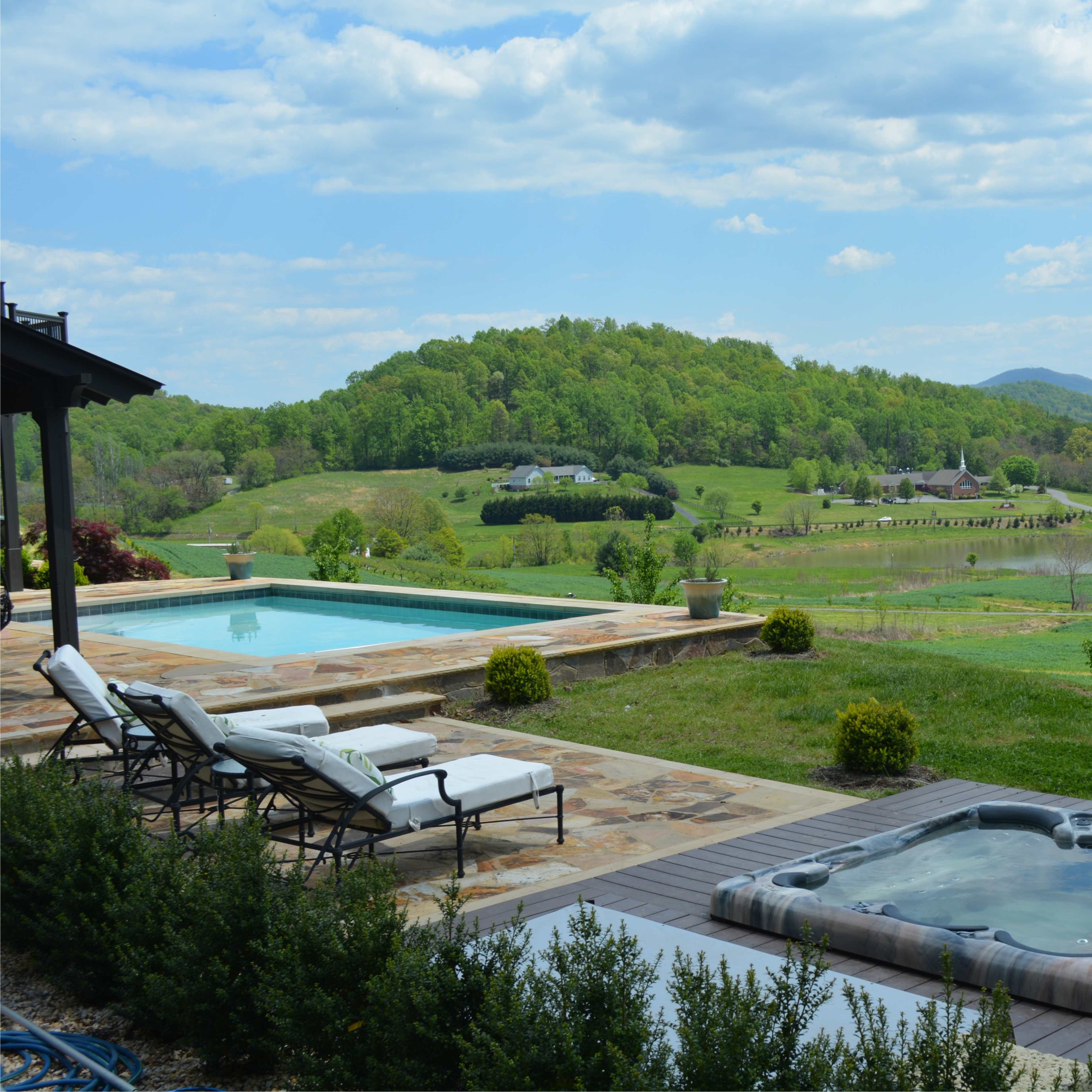 A serene landscape features a swimming pool and hot tub beside a grassy area, with rolling hills and a lake in the background under a partly cloudy sky.