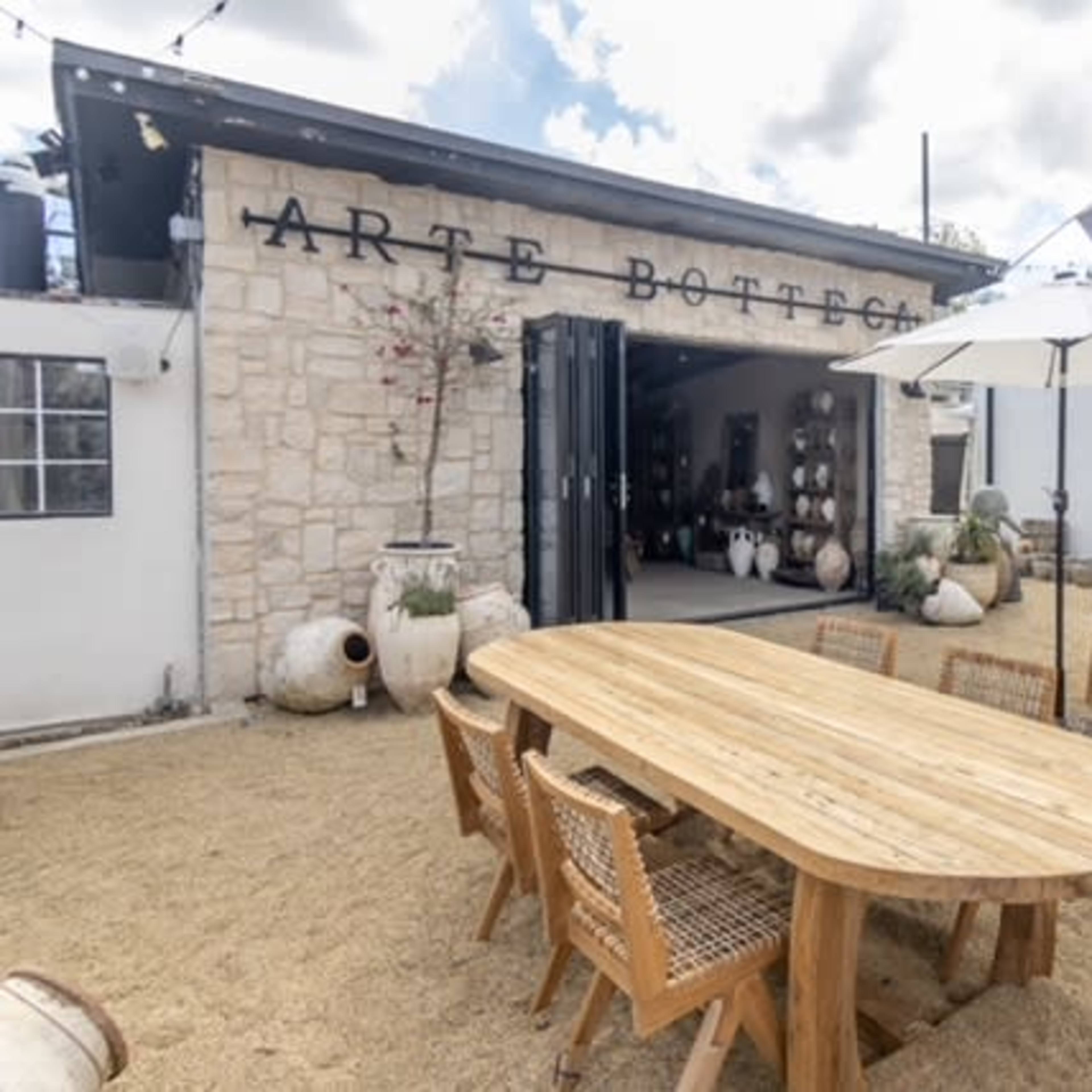 An outdoor dining area featuring a large wooden table surrounded by wicker chairs, set against a backdrop of a building with the sign "Arte Bottega" and various ceramic pots.