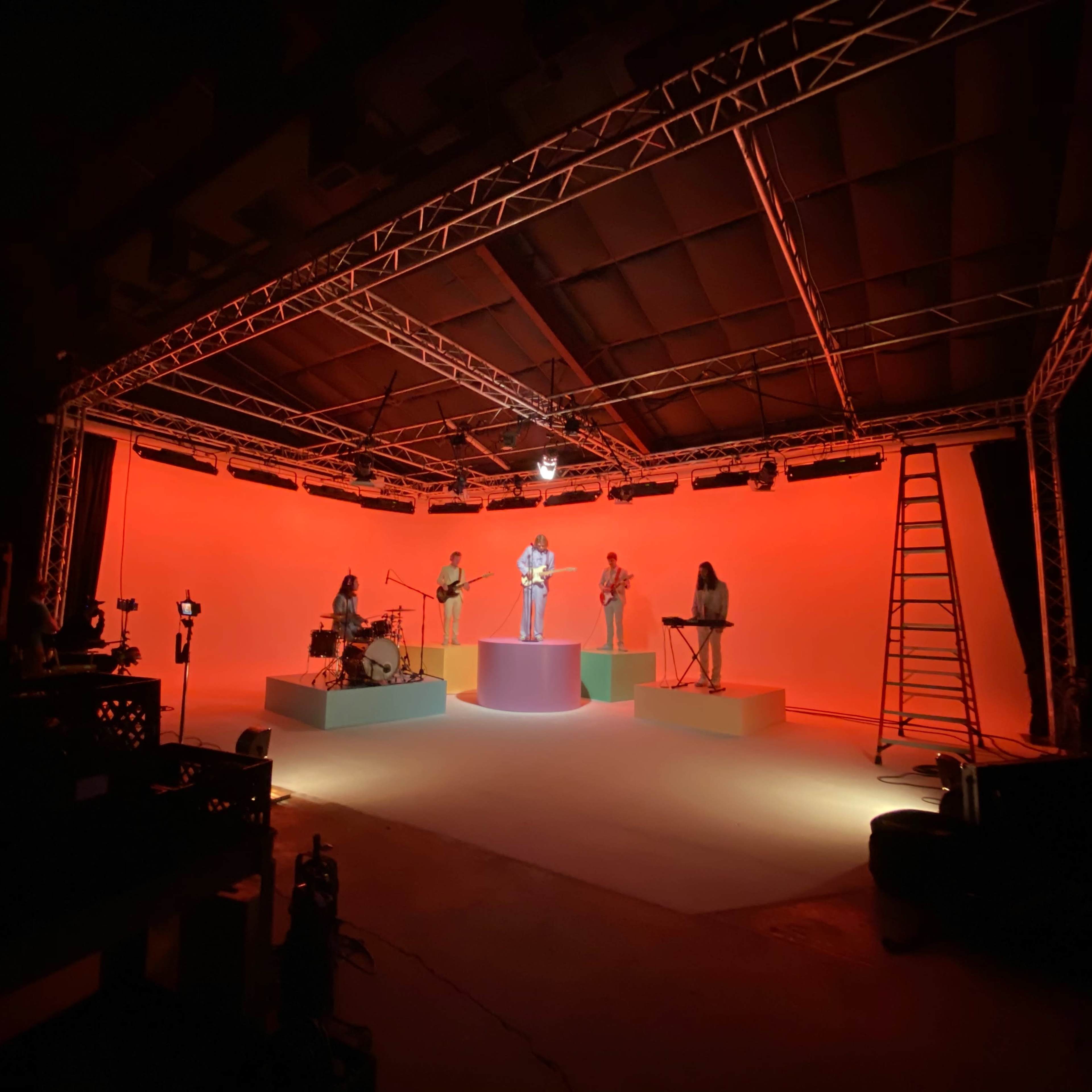 A musical performance is taking place on a brightly lit stage with several musicians standing on colorful platforms against an orange background.