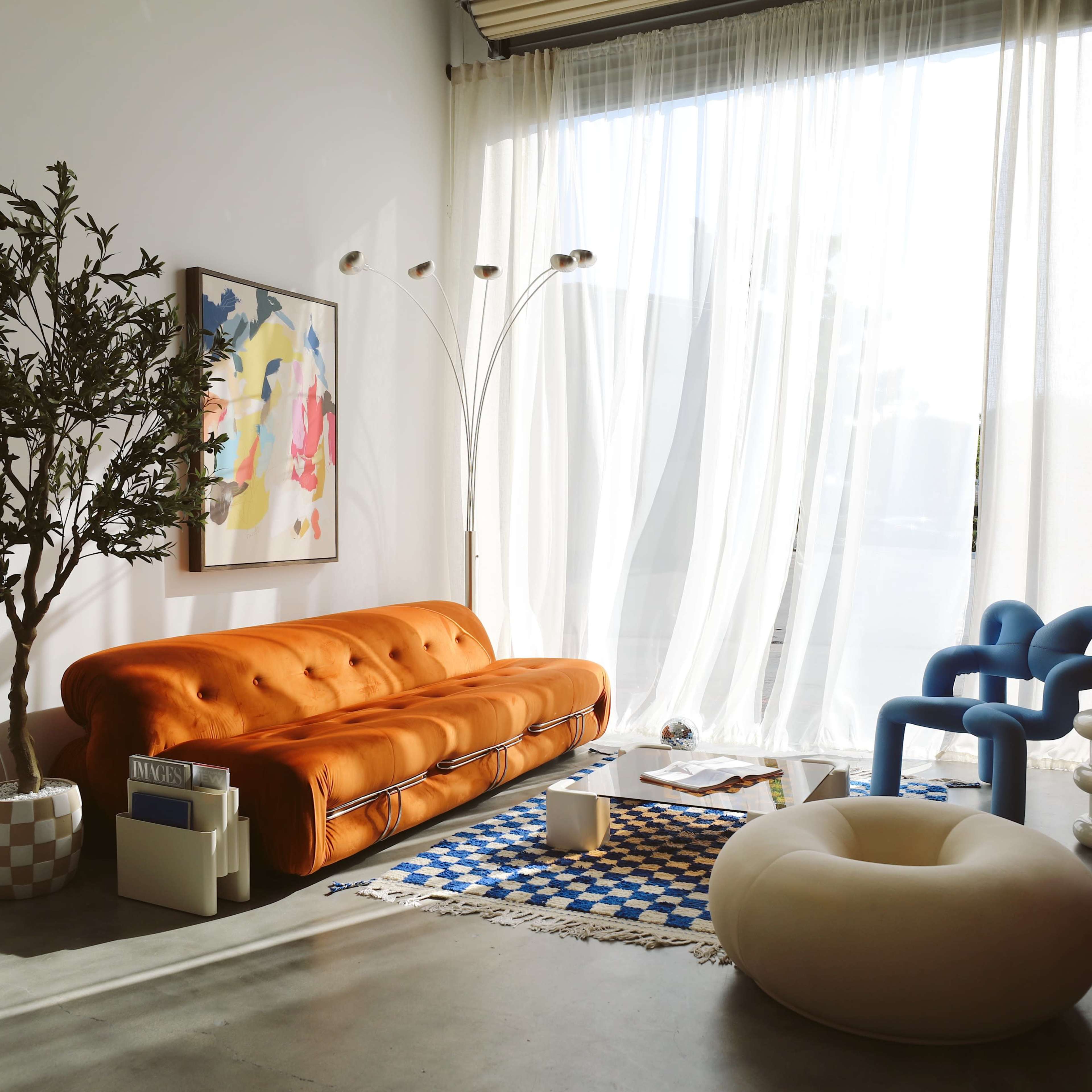 A bright living room featuring an orange sofa, a large window with sheer curtains, a blue chair, and a round white ottoman, with a tree and artwork in the background.