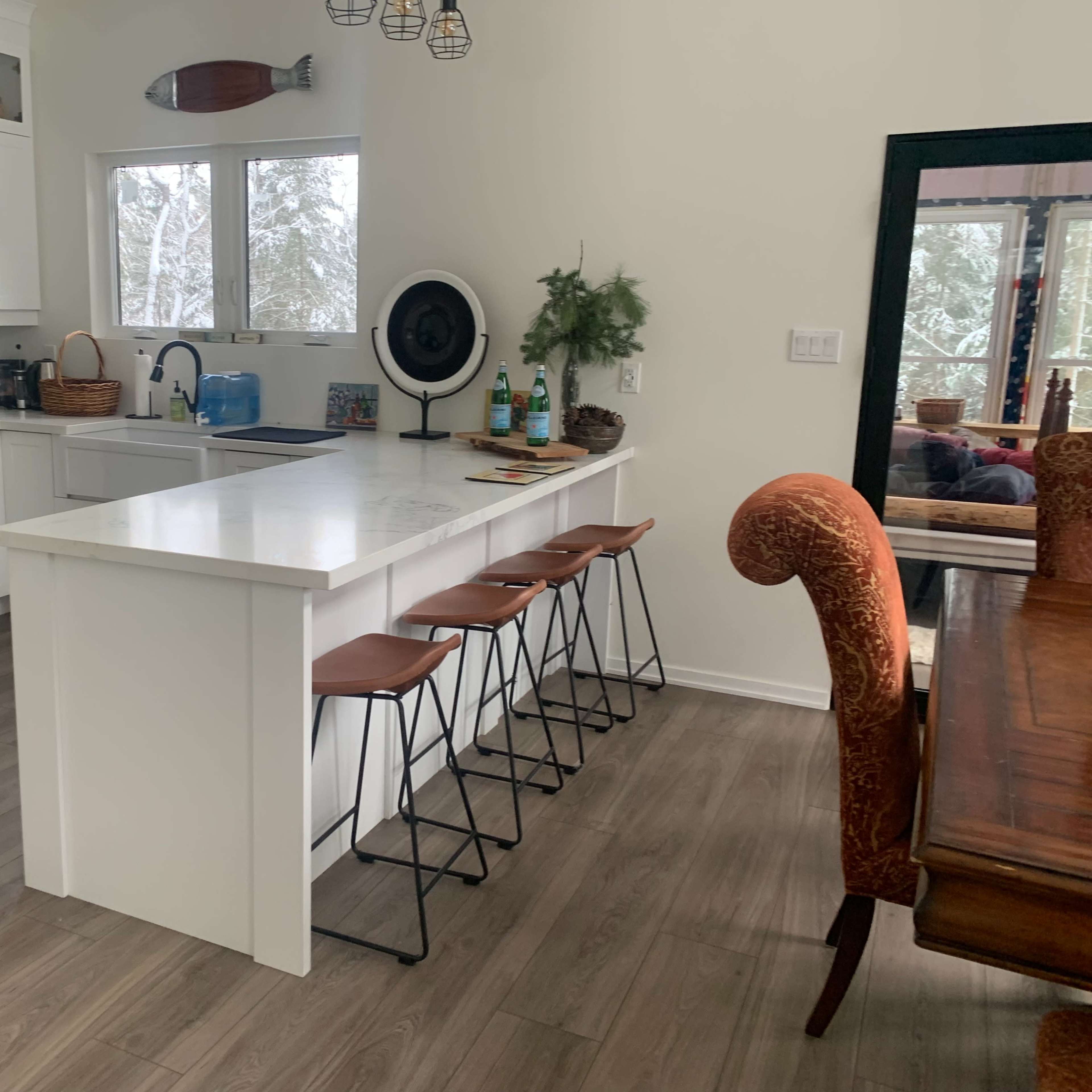 A modern kitchen features a central island with four high stools, stainless steel appliances, and large windows allowing natural light in.