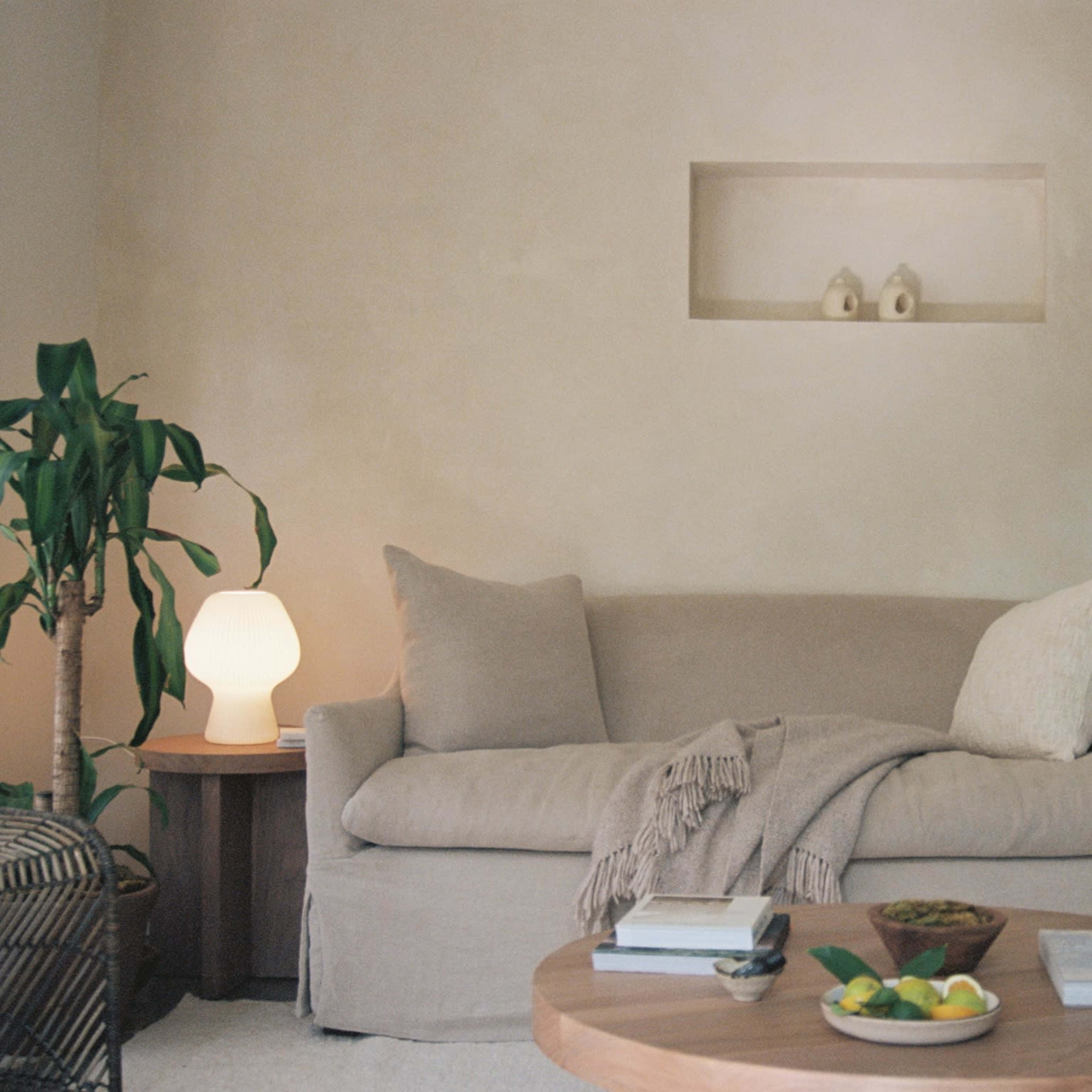 A light-colored sofa with a blanket draped over it sits opposite a wooden coffee table adorned with fruit, surrounded by plants and a lamp.