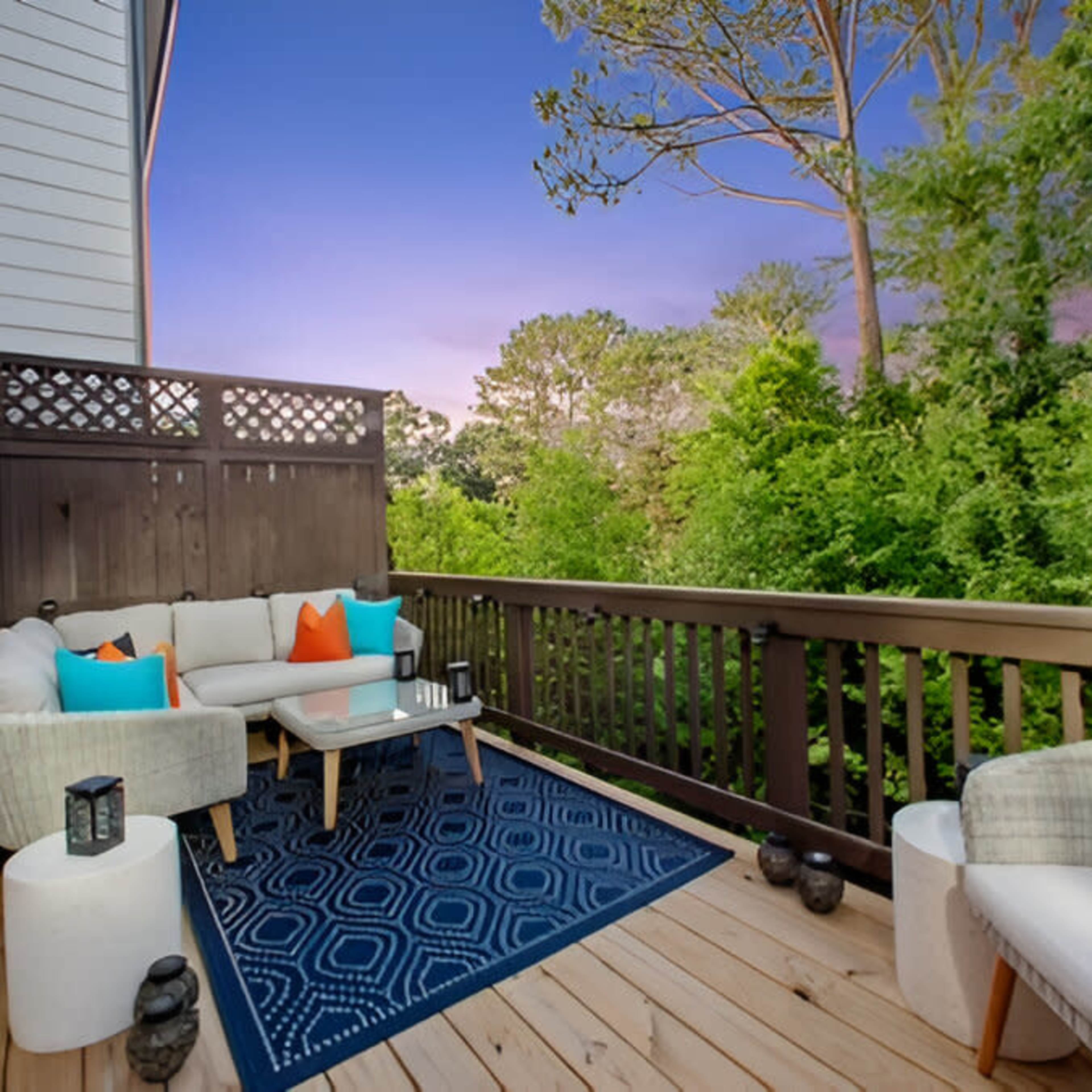 A wooden deck features a seating arrangement with colorful cushions, a coffee table, and decorative lanterns, surrounded by lush greenery.