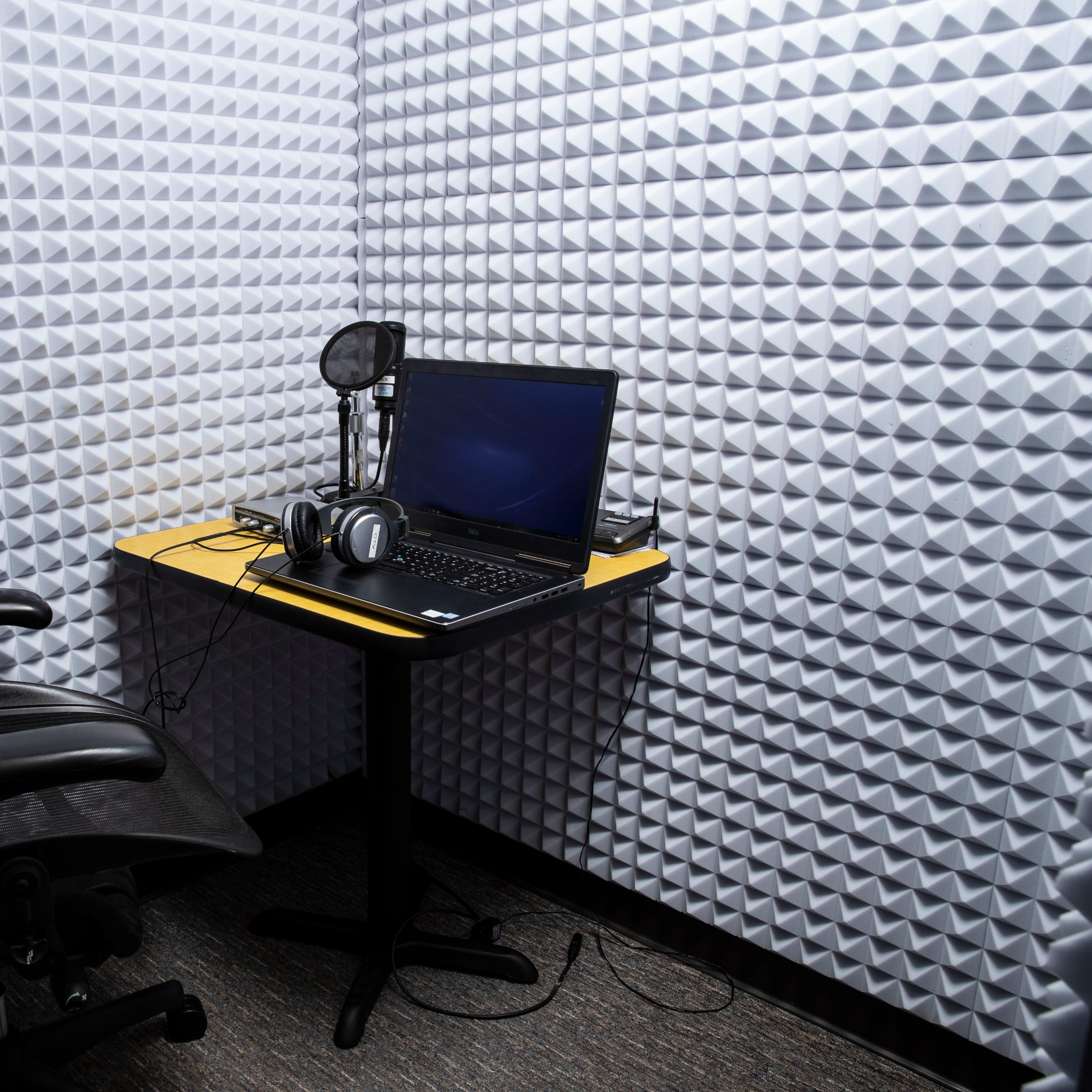 A small recording studio features a desk with a laptop, headphones, and a microphone, surrounded by soundproof foam panels.