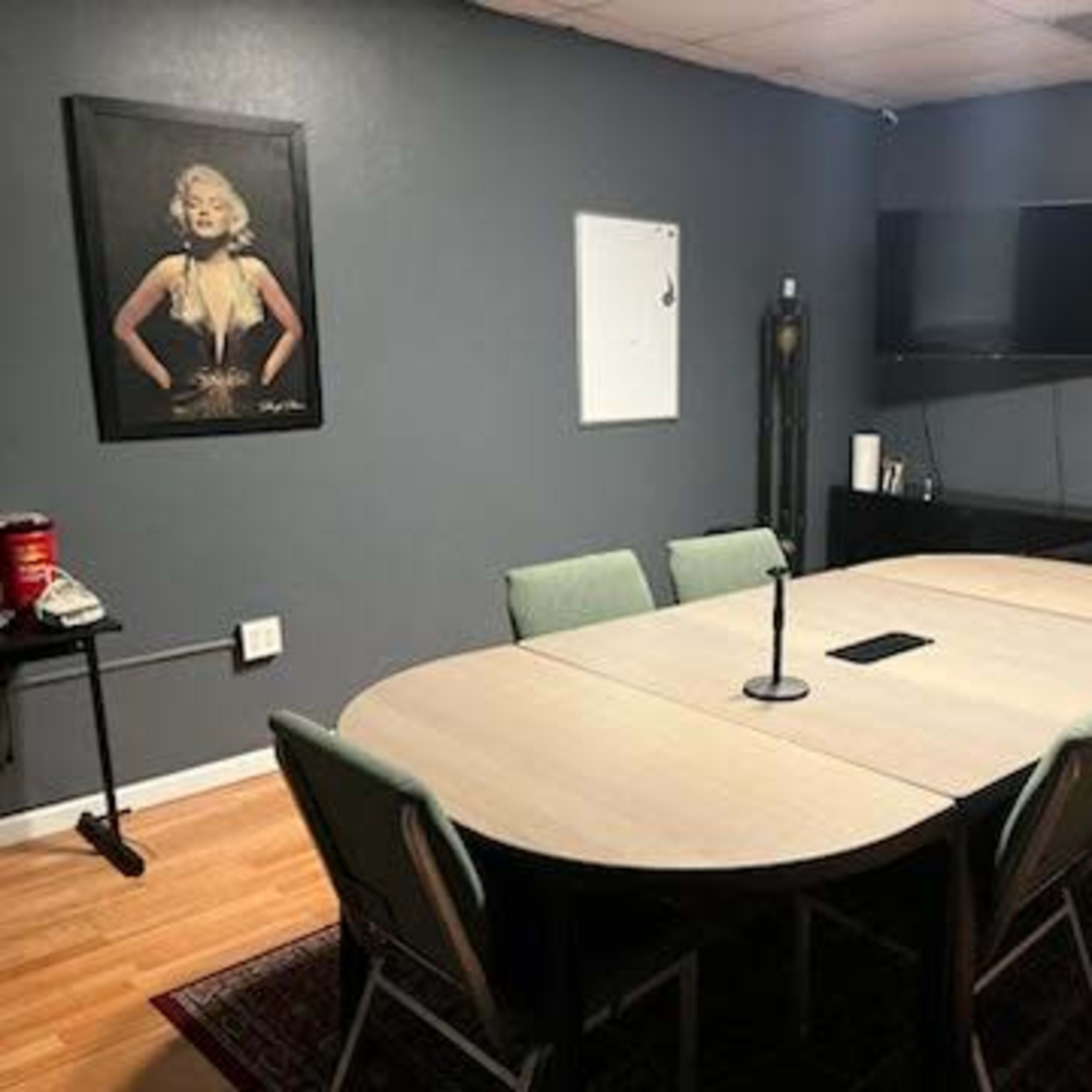 A conference room with a large oval table surrounded by eight green chairs, a wall-mounted TV, and a framed poster on the wall.