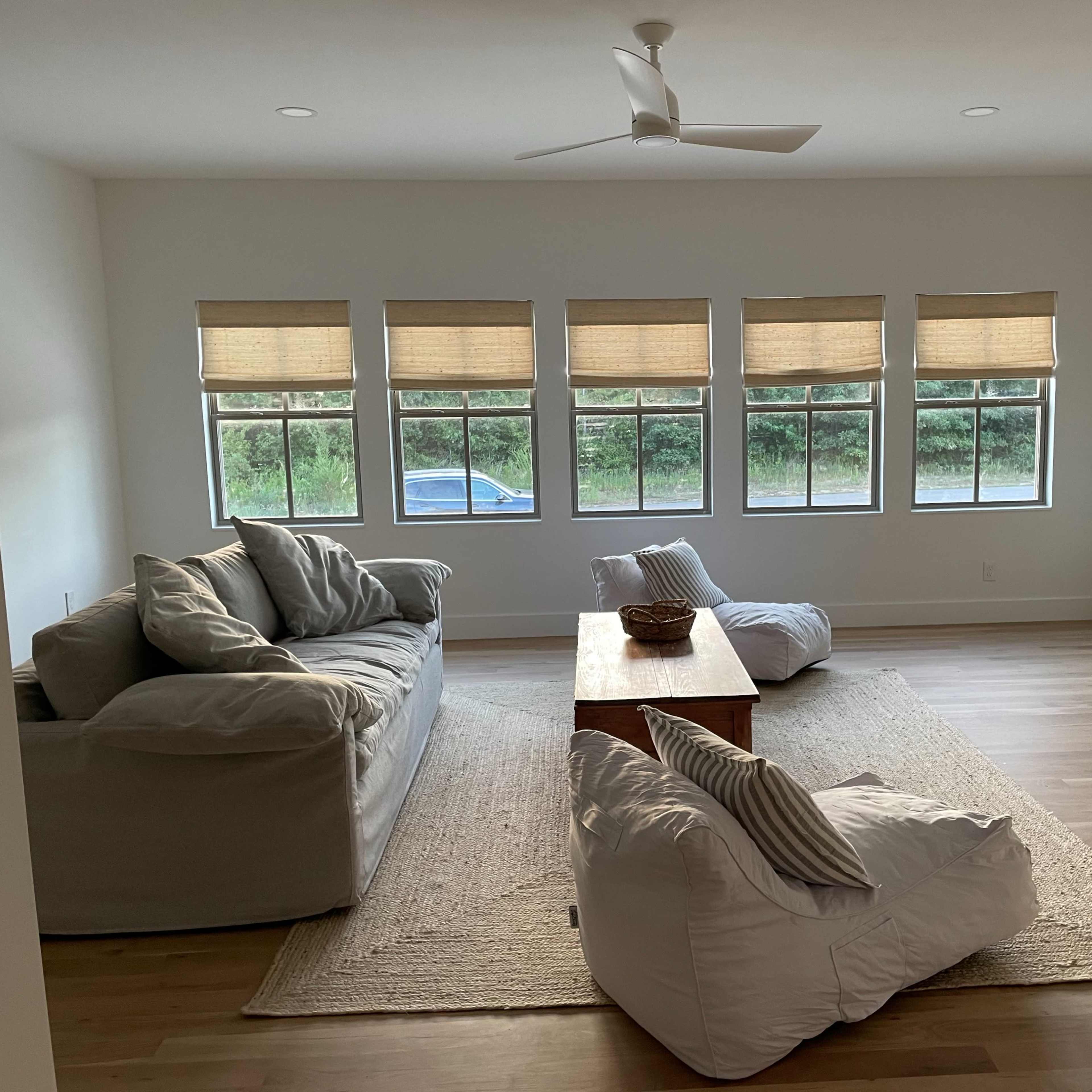 A spacious living room features two sofas, a wooden coffee table, and bean bag chairs, with large windows allowing natural light to illuminate the space.