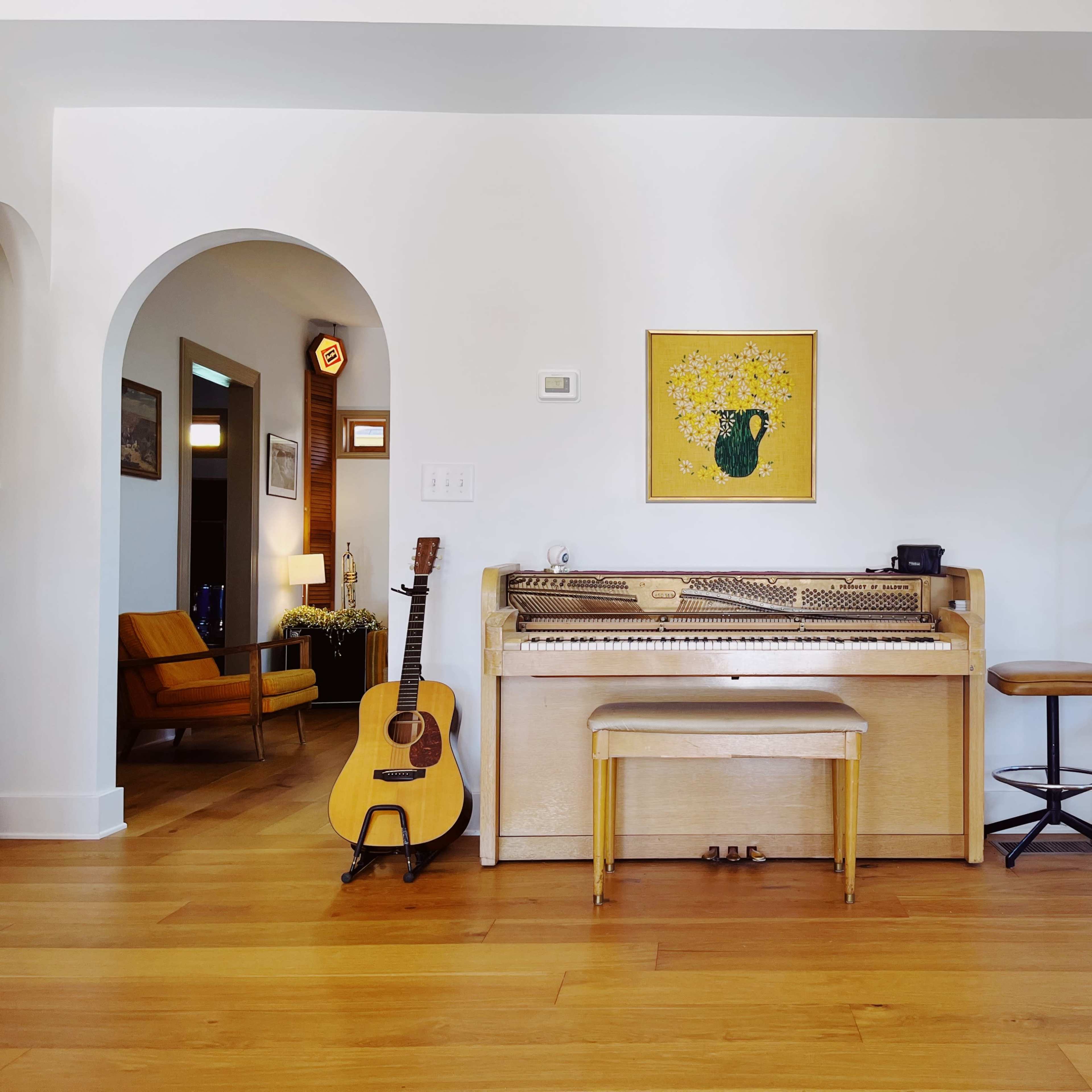 A light-colored piano with a bench in front of it, accompanied by an acoustic guitar, set against a bright, minimalistic interior featuring an archway and a small bookshelf.