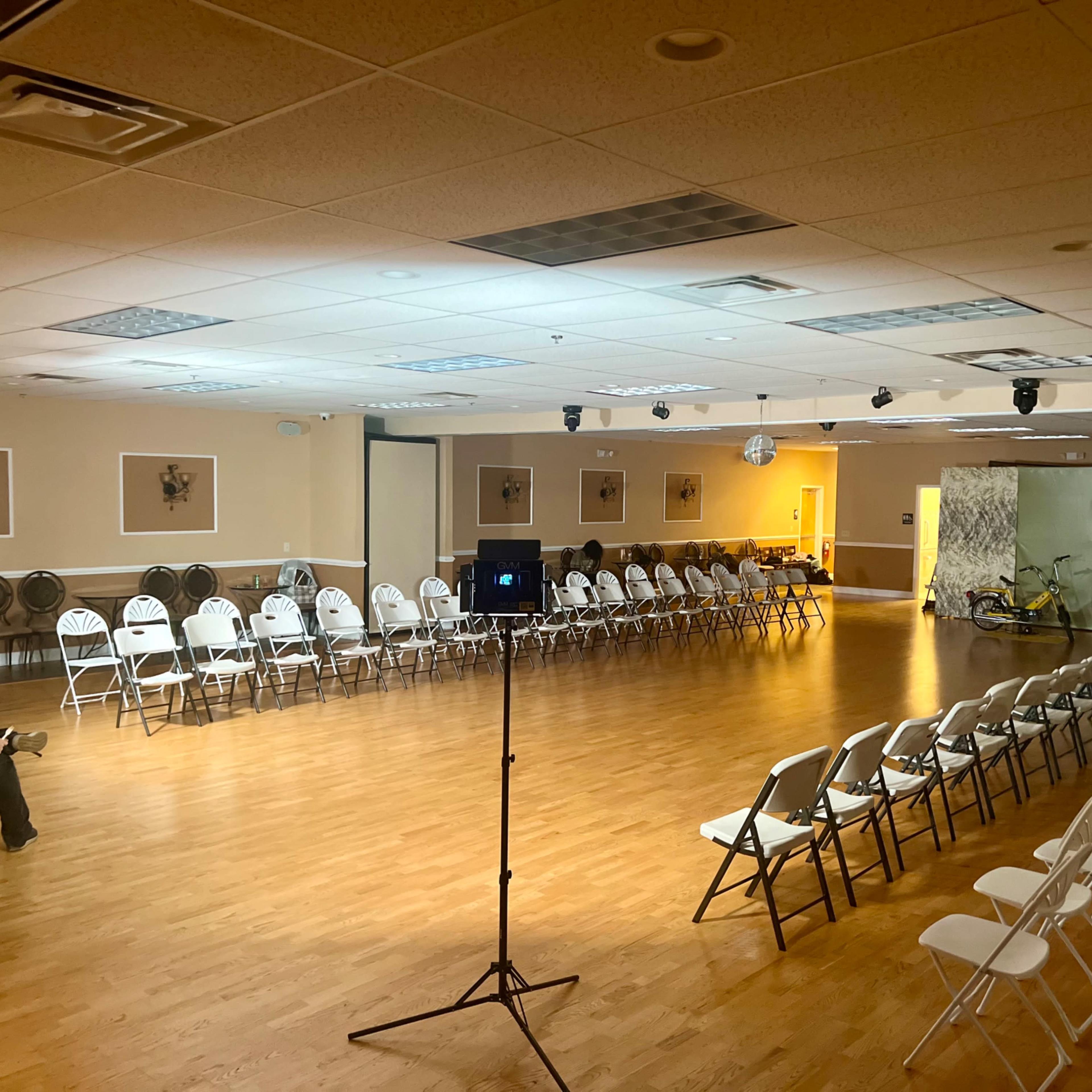 A spacious event room with rows of white chairs arranged in two parallel lines, and a small stage set at one end.