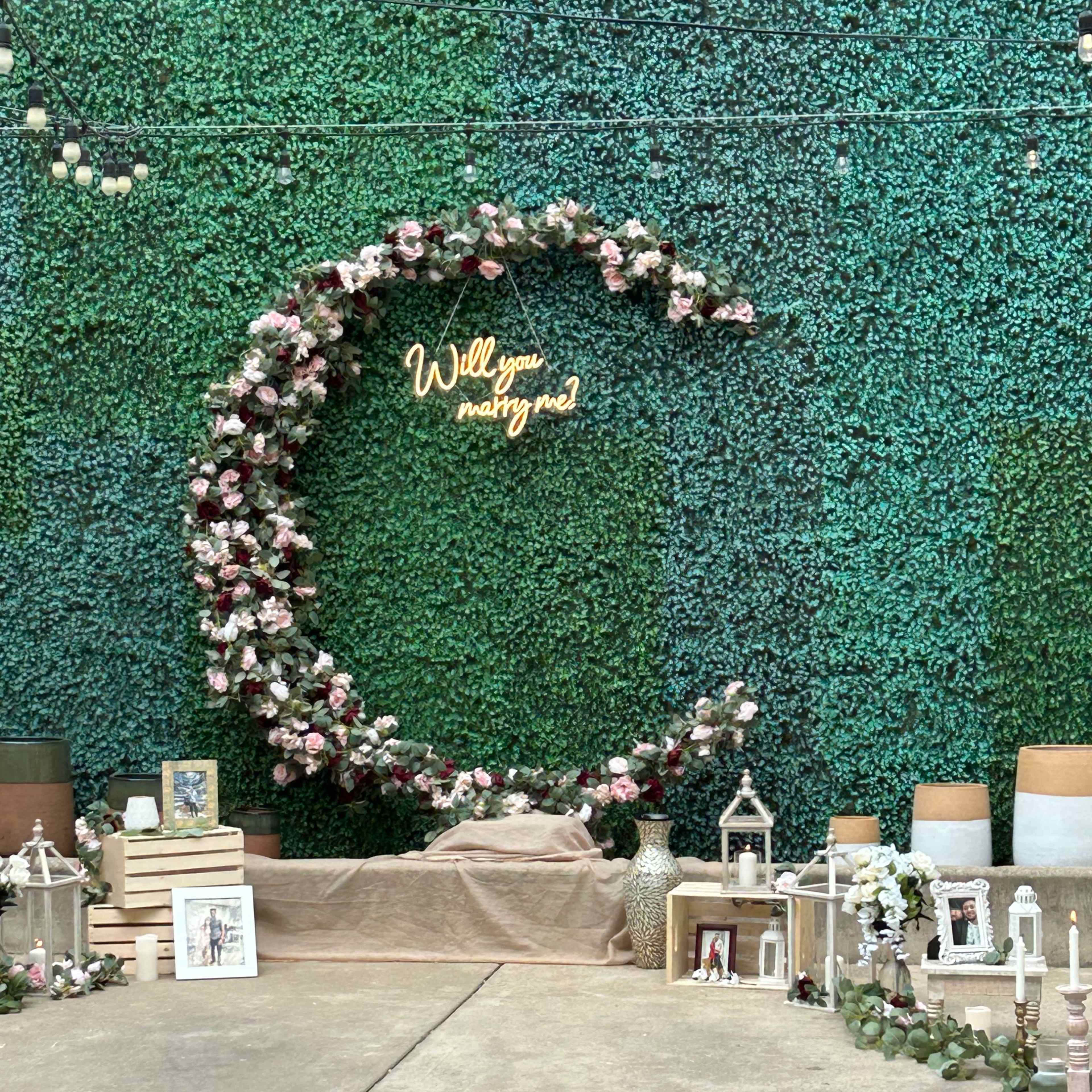 A decorative setup for a marriage proposal, featuring a floral arch with the phrase "Will you marry me?" illuminated in lights against a green backdrop.