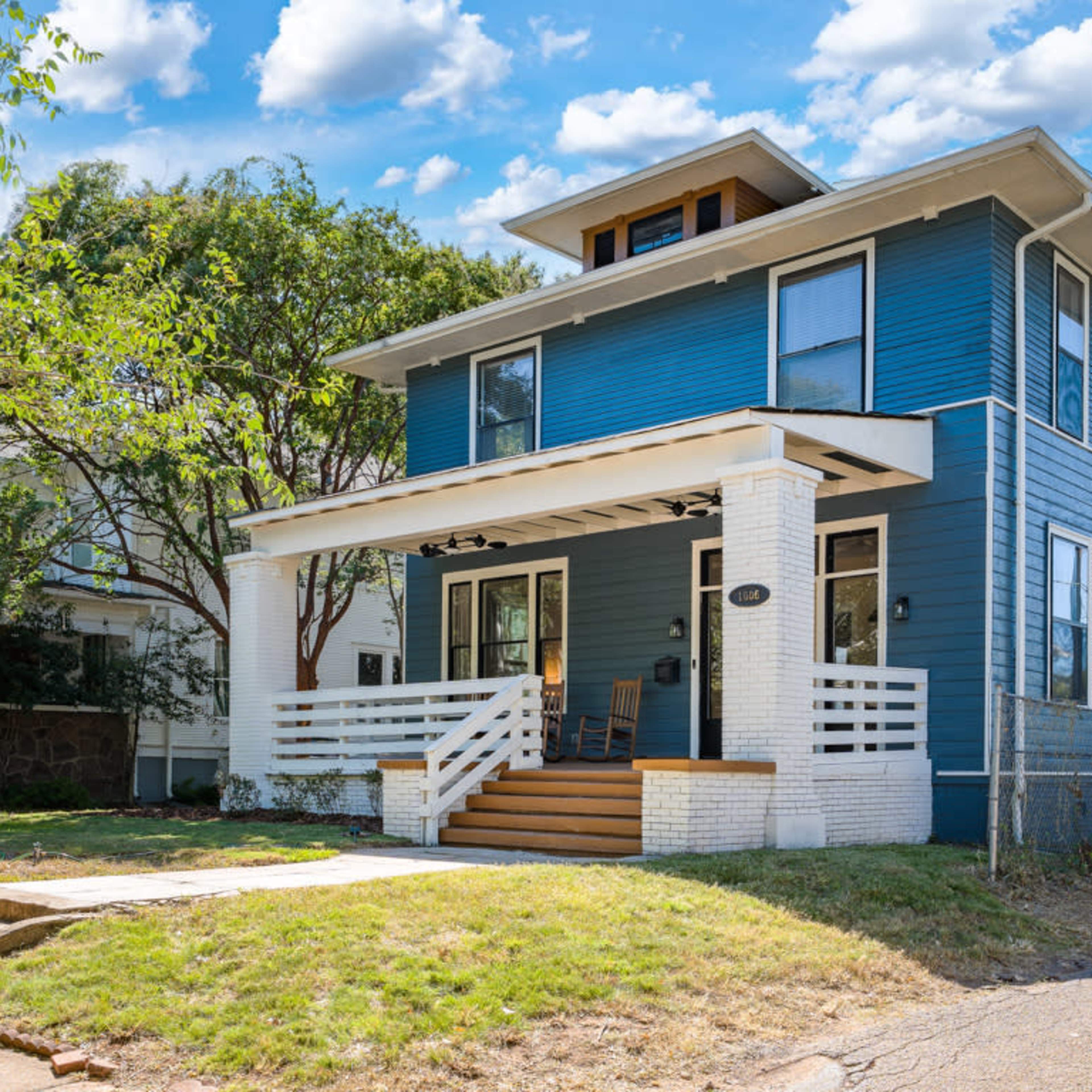 A three-story blue house with a front porch, white trim, and a tree-filled lawn on a quiet street.