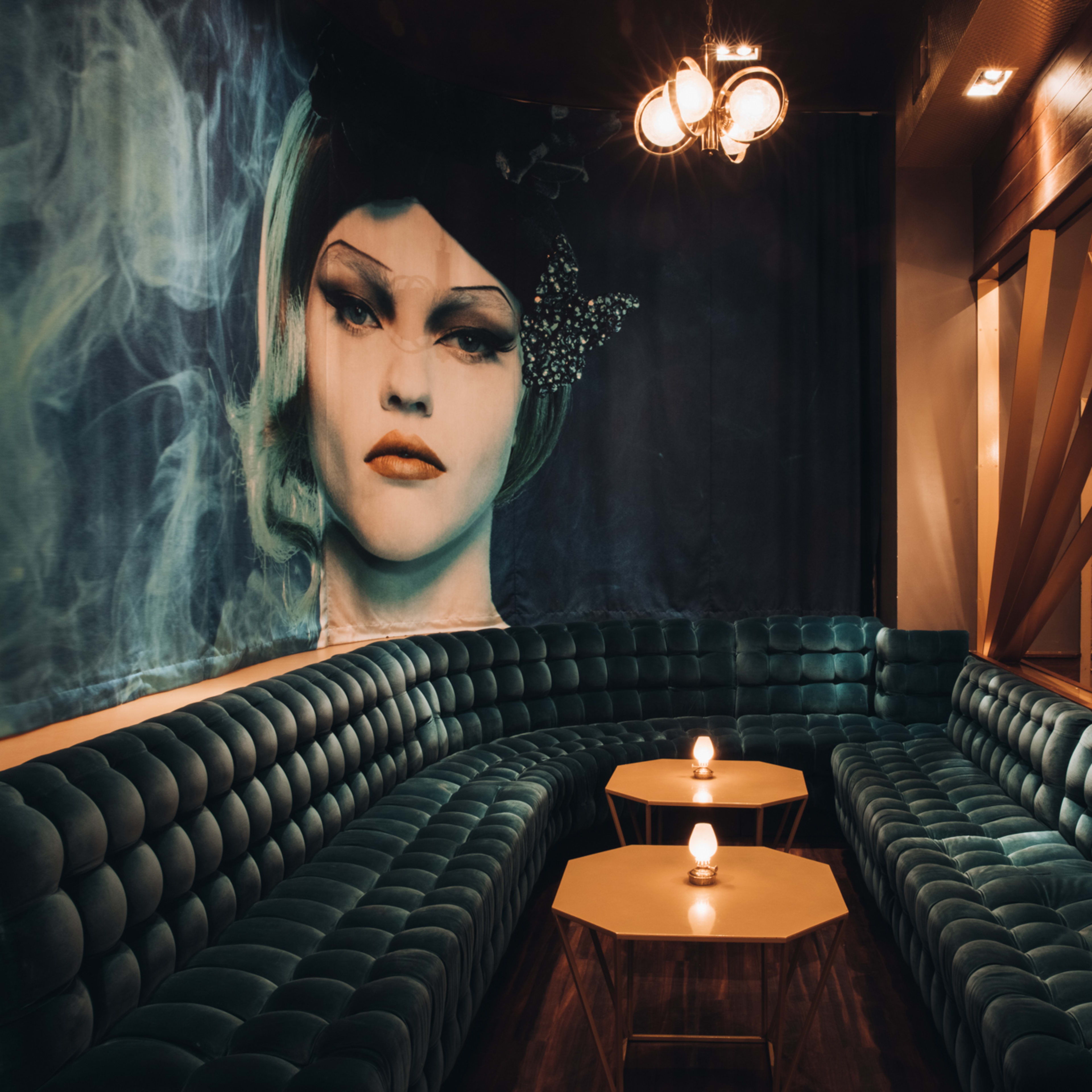 A plush, curved green velvet sofa is positioned against a wall featuring a large mural of a woman's face, with two small tables in front holding lit candles.