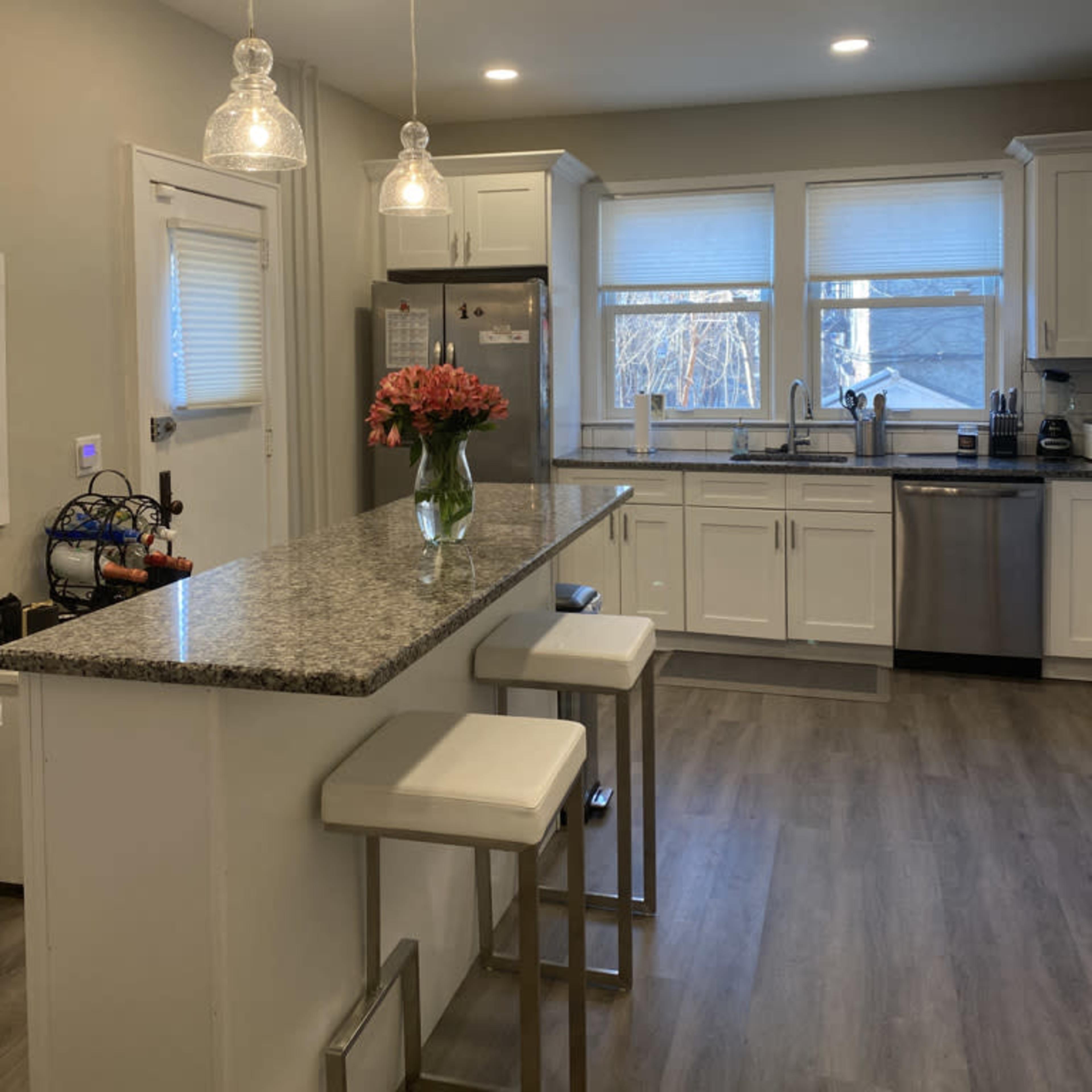 A modern kitchen features white cabinetry, a granite countertop island with seating, and stainless steel appliances, illuminated by pendant lighting.