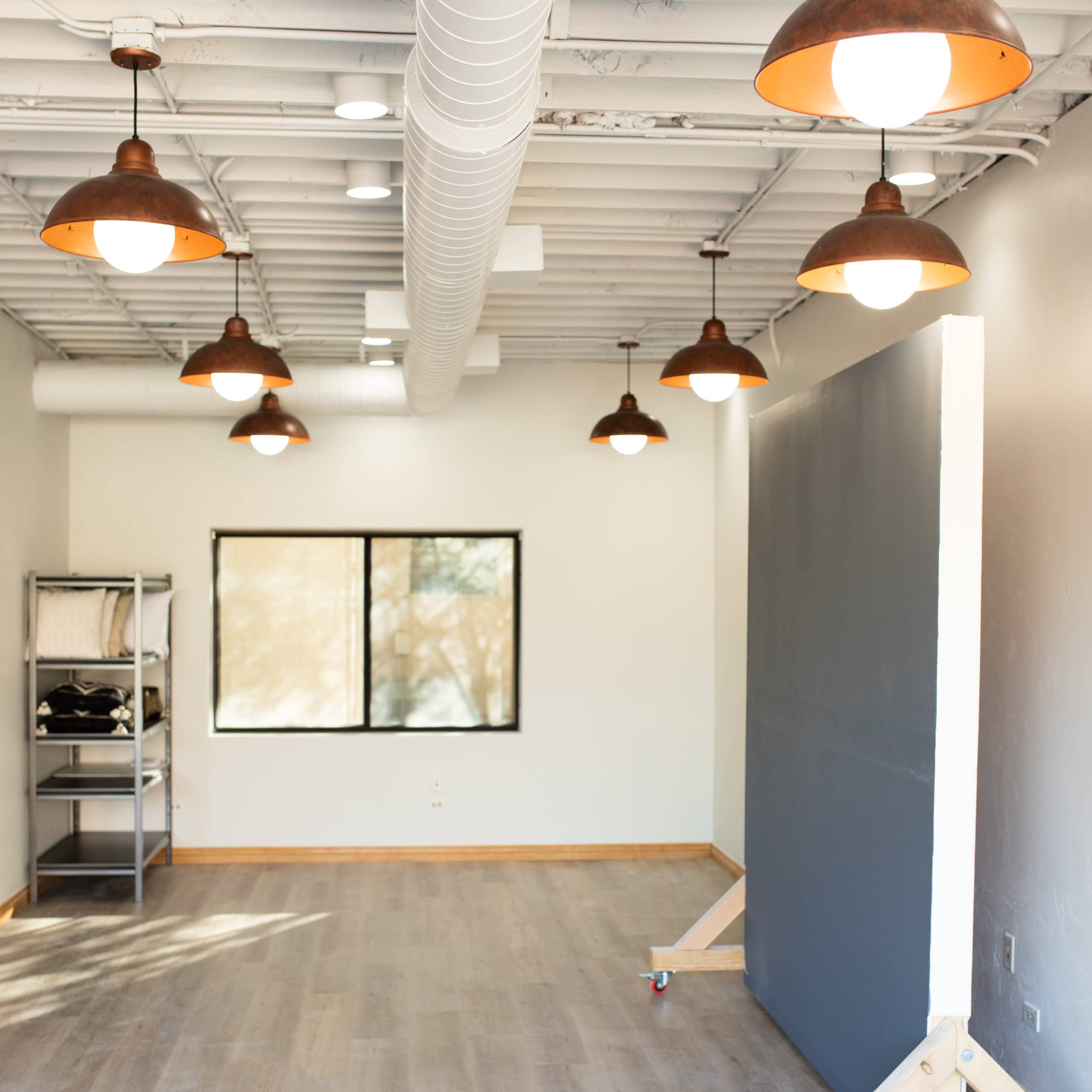 A spacious room with wooden flooring, featuring overhead industrial-style pendant lights and a large, movable gray panel on a wheeled stand.