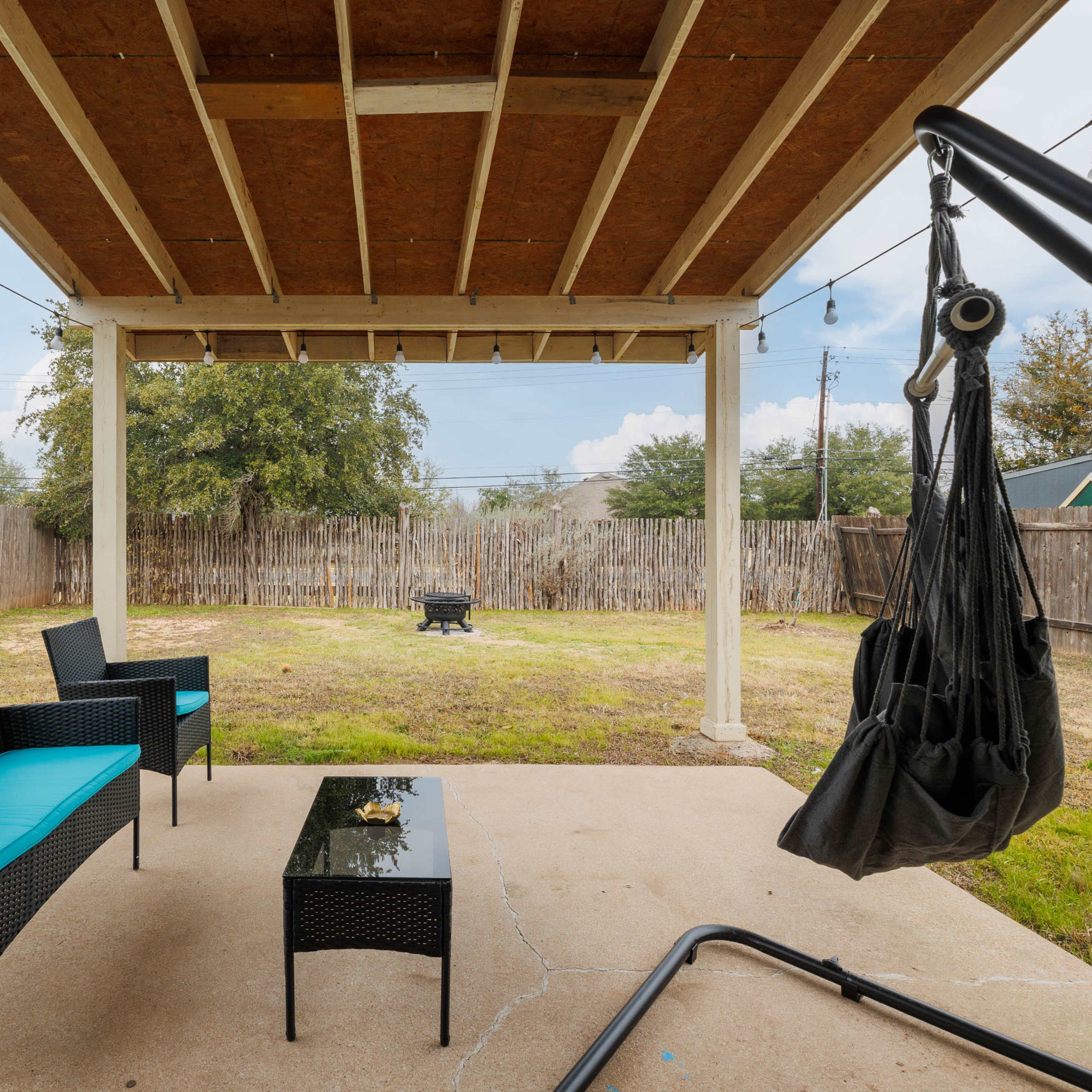 A covered patio area features black wicker furniture with teal cushions, a glass-top coffee table, and a hanging chair, overlooking a spacious grassy yard enclosed by a wooden fence.