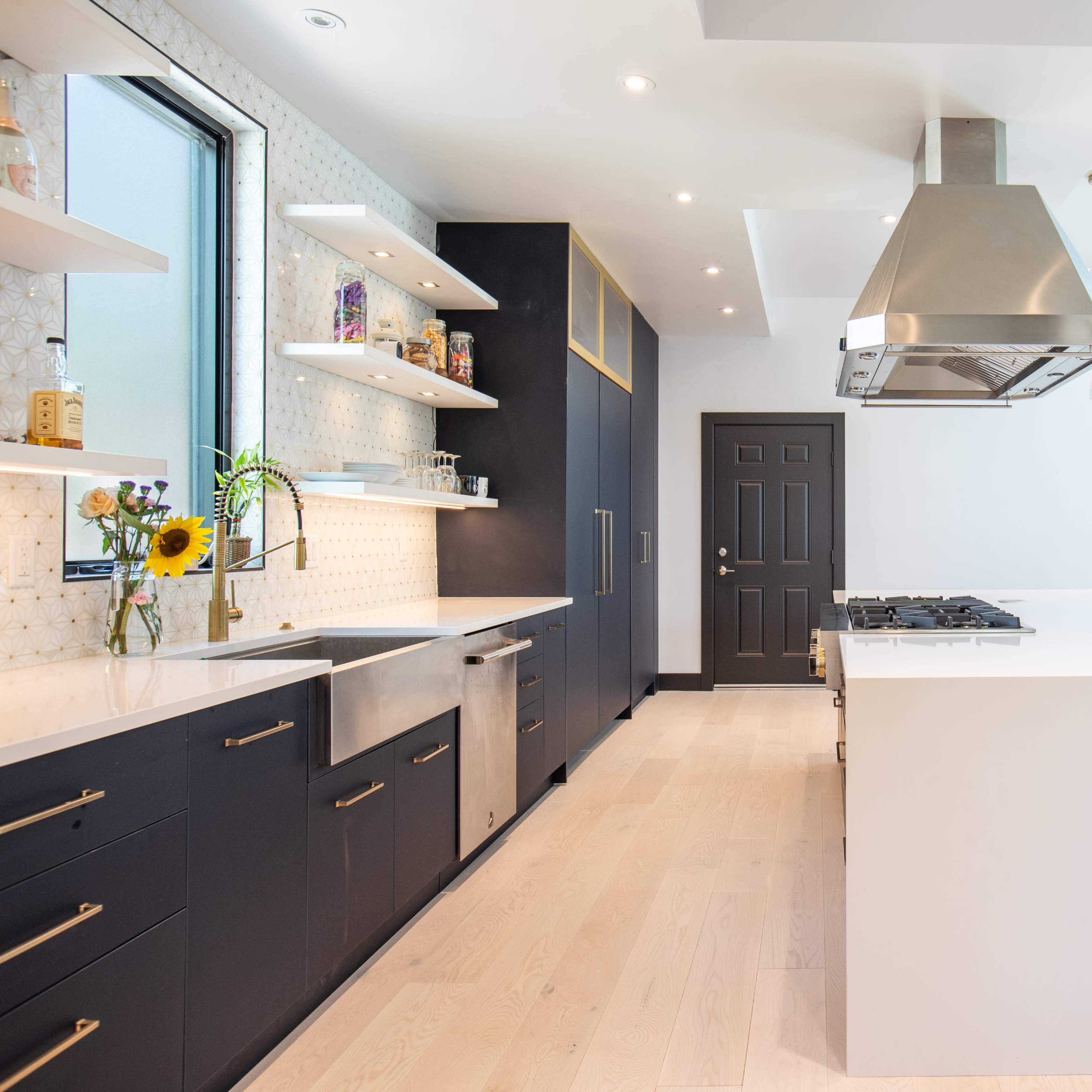A modern kitchen features white countertops, dark cabinets, a central island with a stovetop, and large windows allowing natural light.
