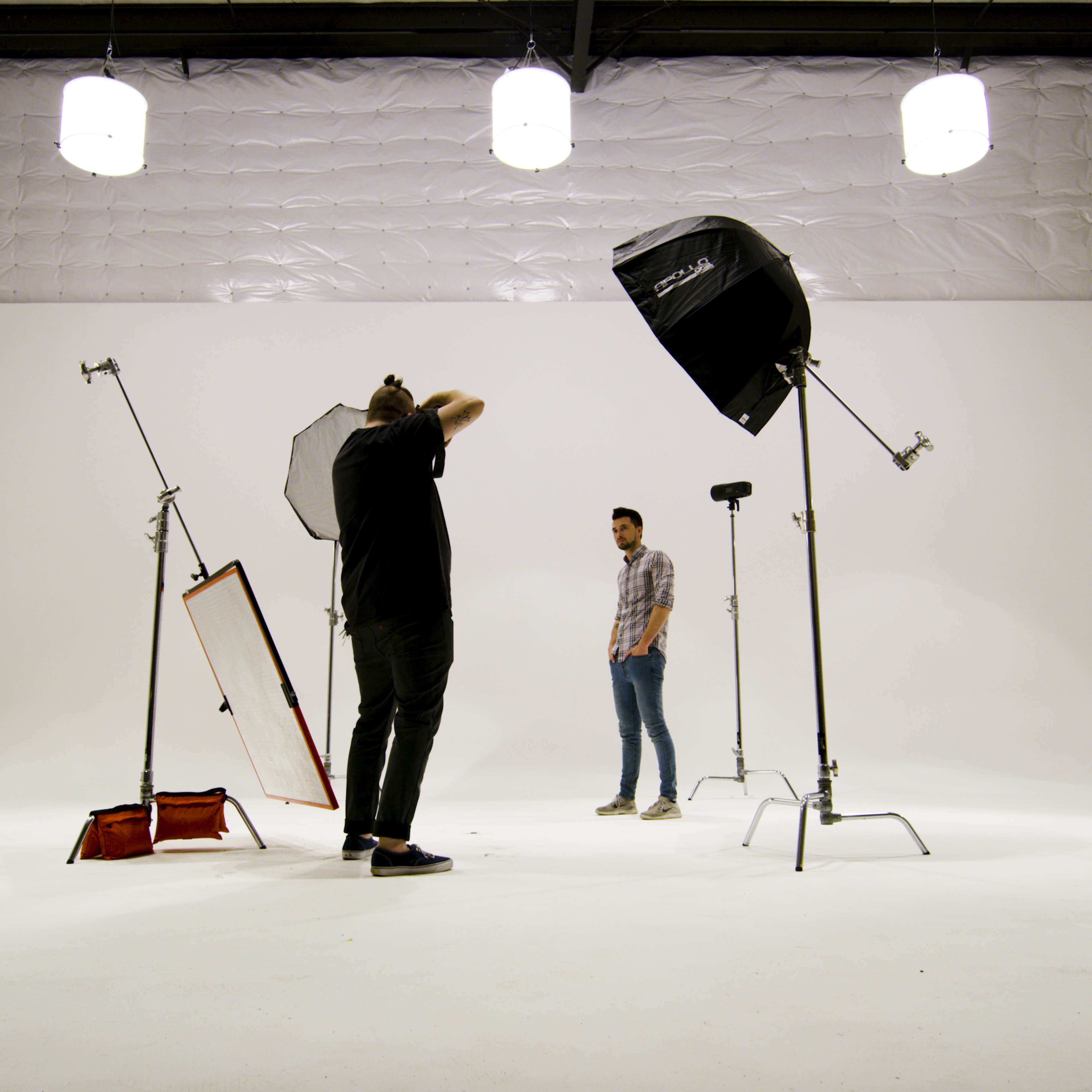 A photographer is adjusting equipment in a spacious, well-lit studio while a model stands in front of a white backdrop.