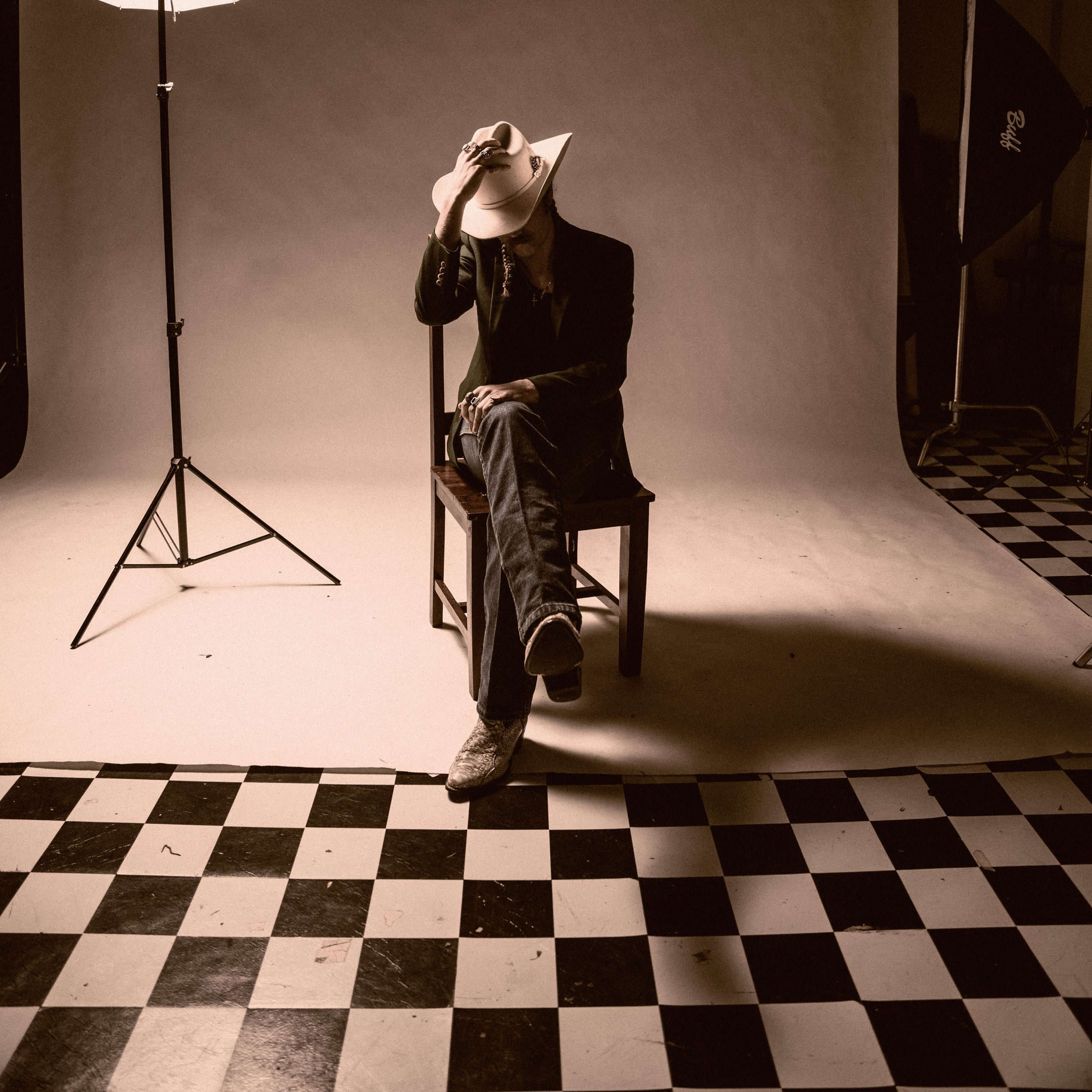 A person wearing a cowboy hat sits on a wooden chair in a studio with black and white checkered flooring, illuminated by a large softbox light.