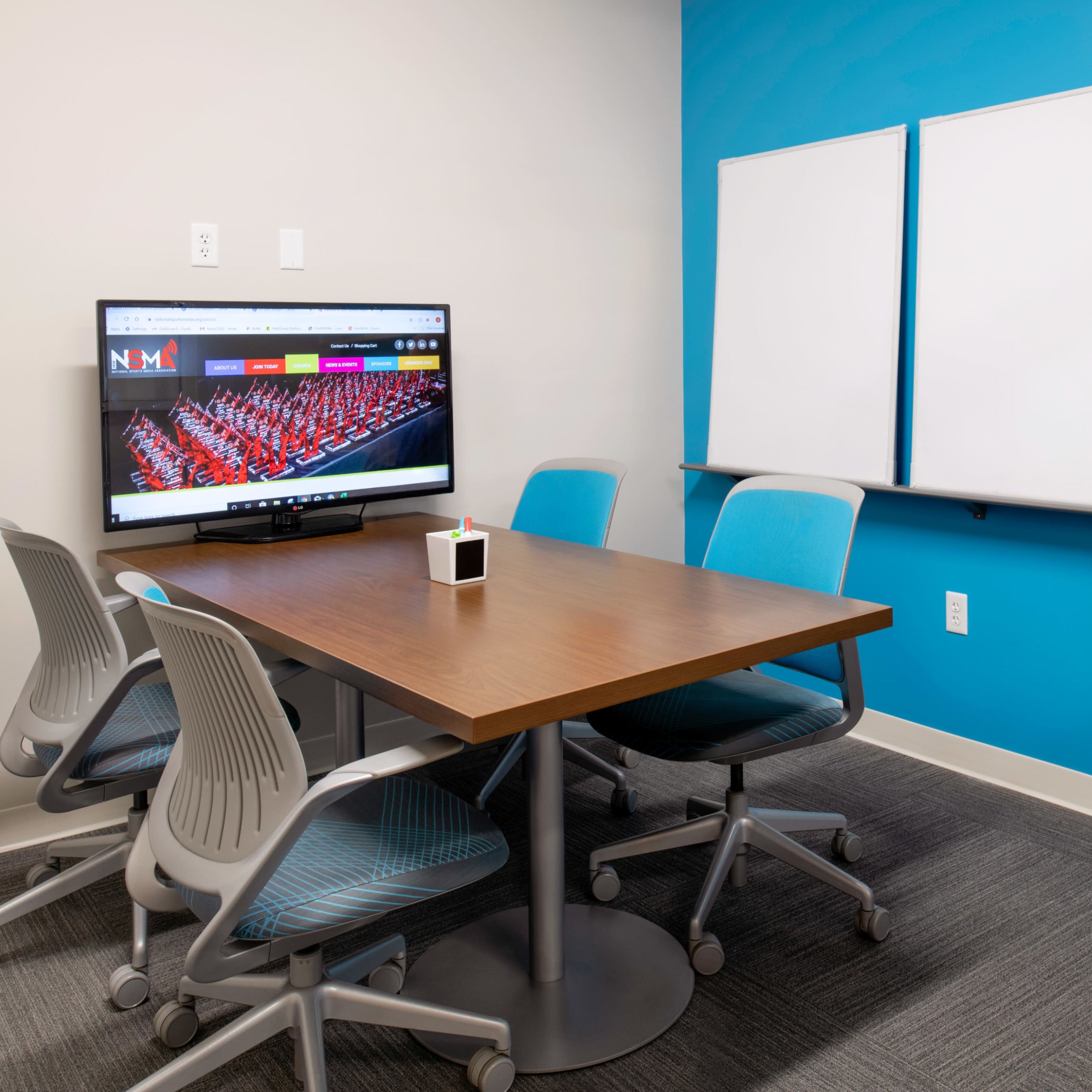 A small meeting room with a wooden table, four chairs, a television displaying a website, and a pair of whiteboards on the wall.