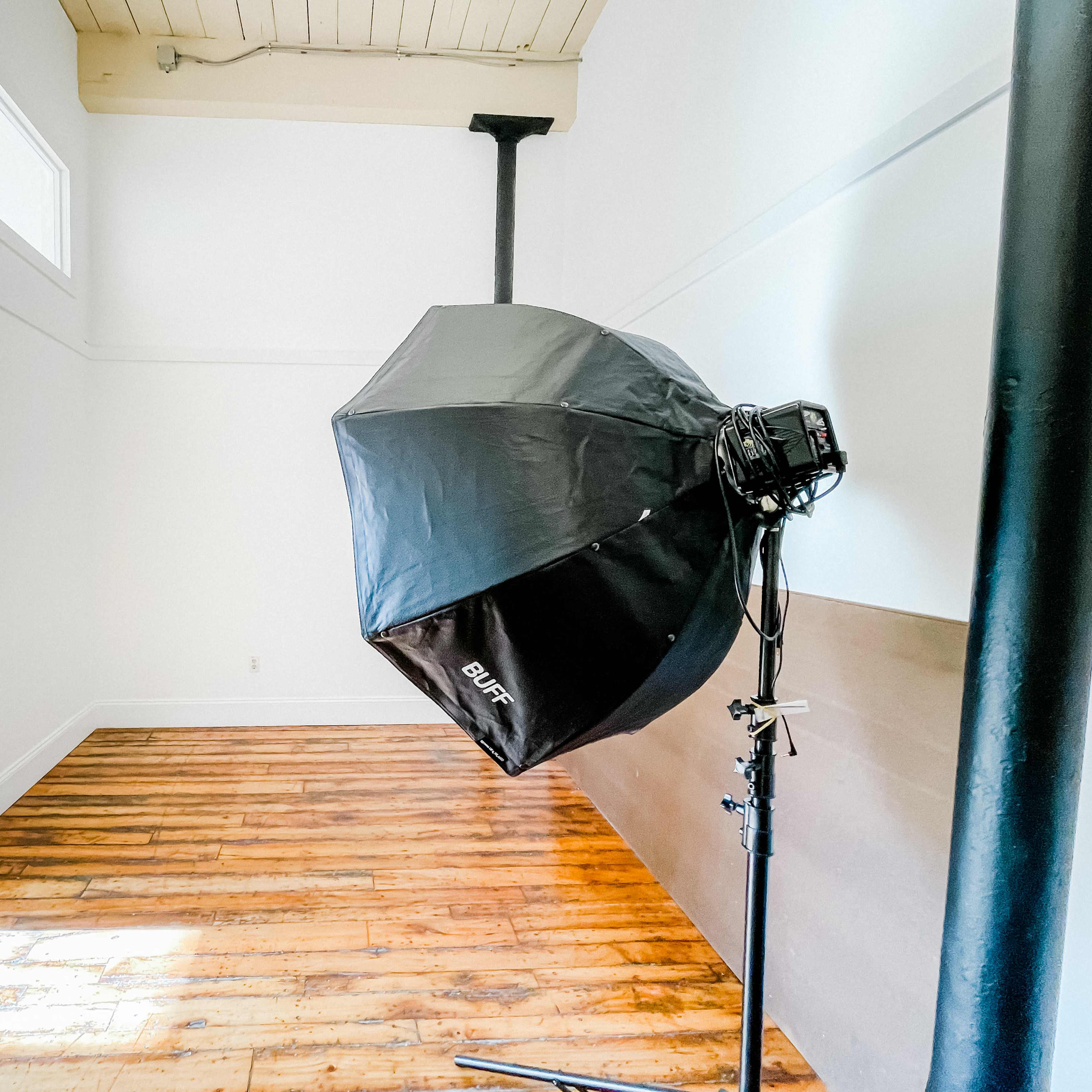 A large softbox light stands on a stand in an empty room with wooden floors and white walls.