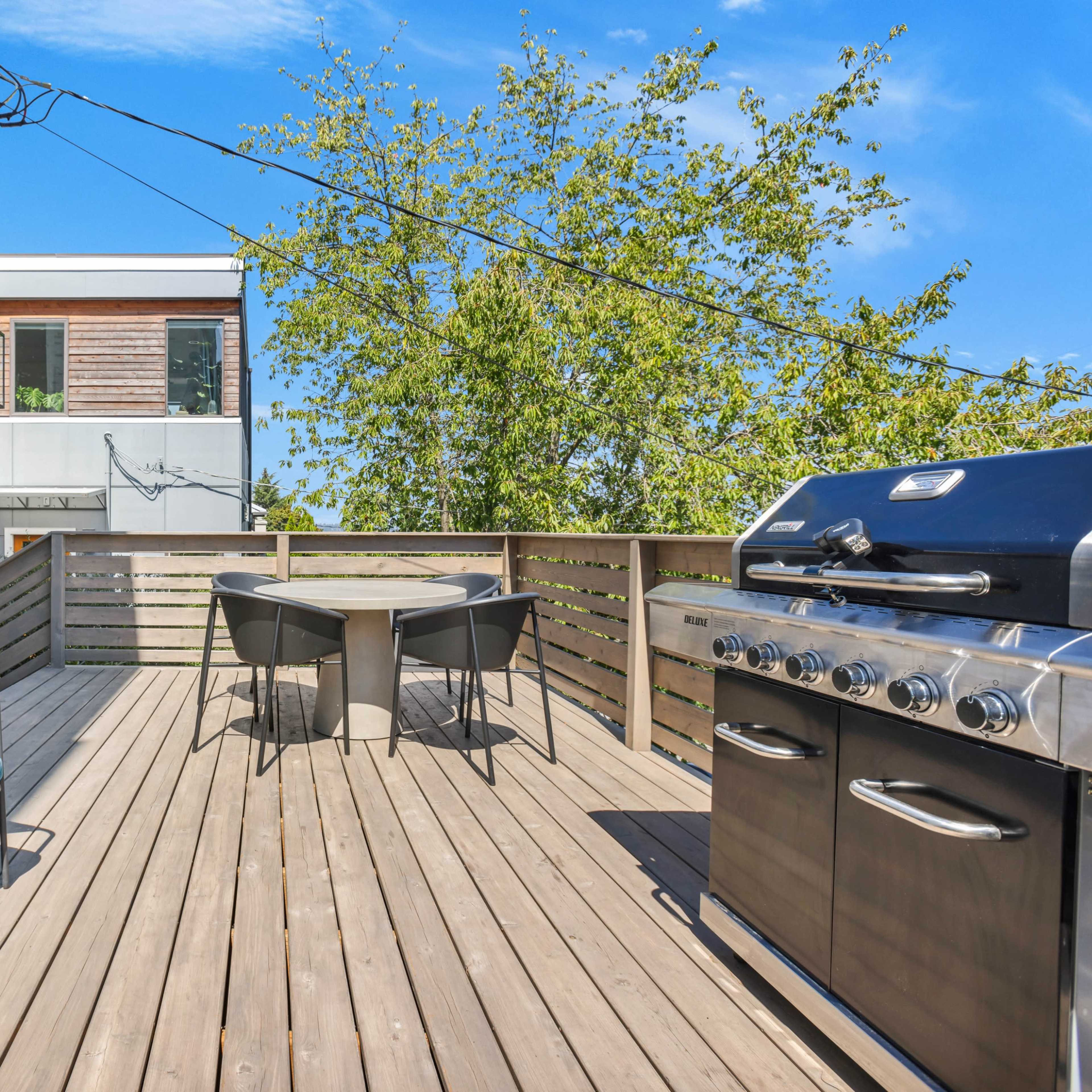 A deck features a stainless steel grill, a circular table with chairs, and views of surrounding trees.