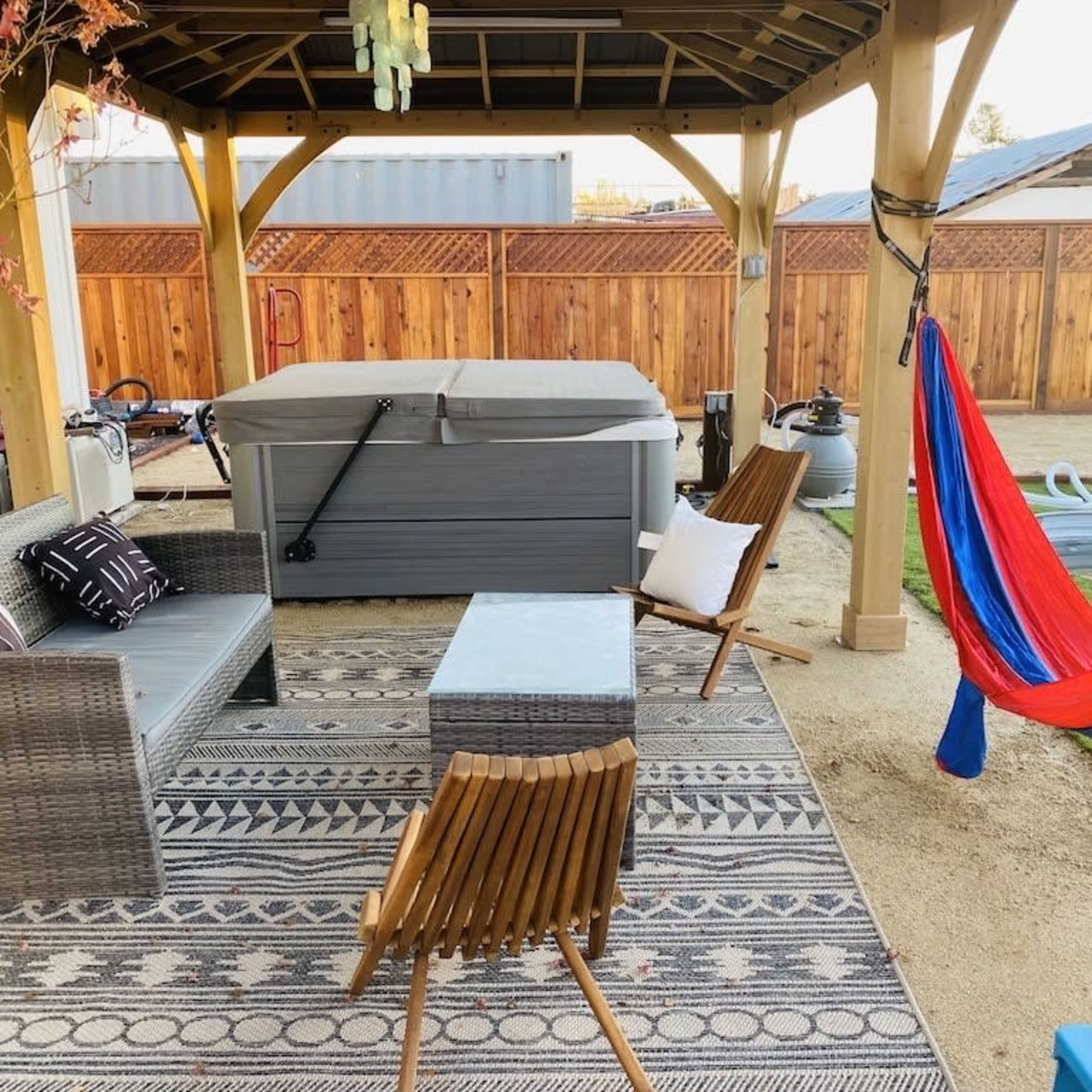 A covered outdoor patio area with a hot tub, seating, a patterned rug, and a colorful hammock.