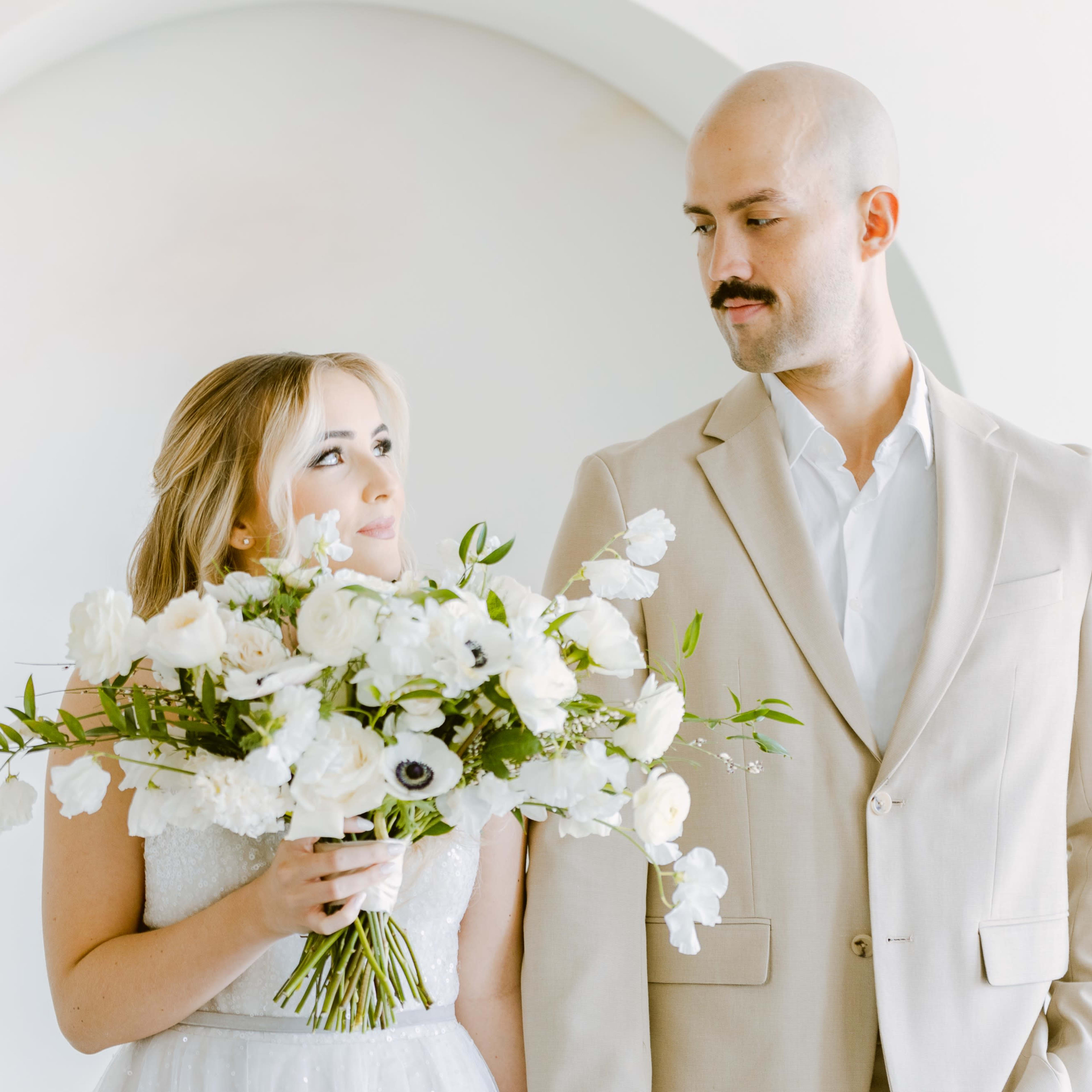 A couple stands side by side, with the woman holding a large bouquet of white flowers and the man dressed in a light suit.