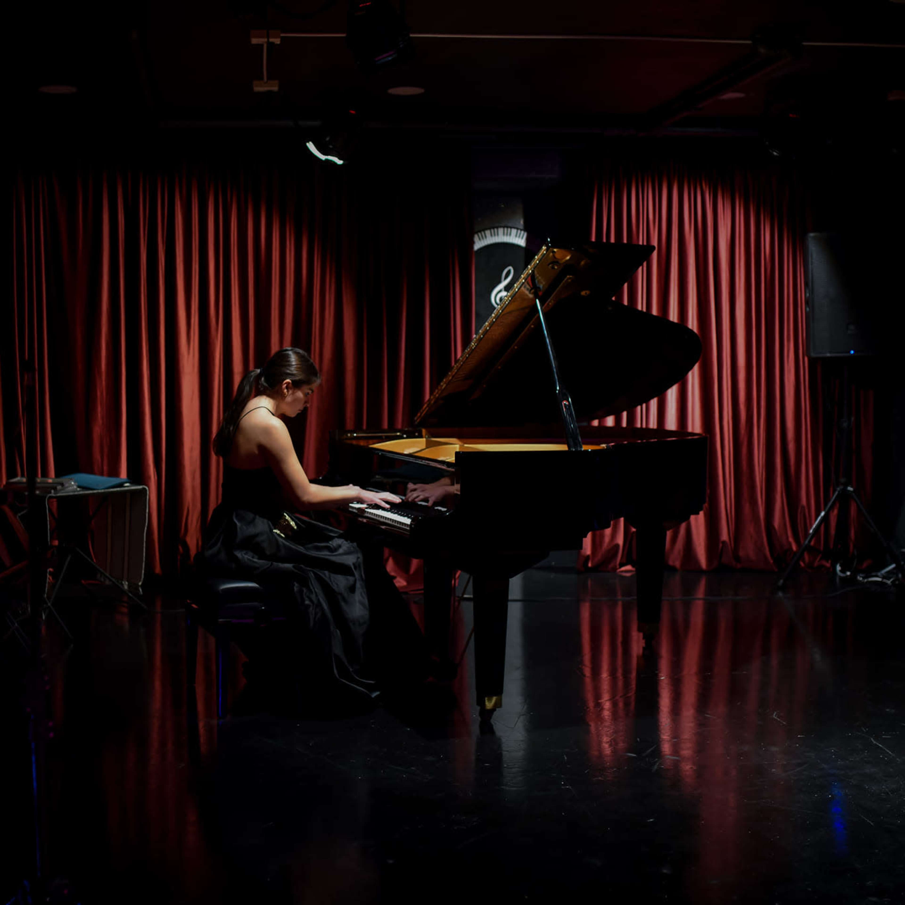 A woman in a black dress plays a grand piano on a dimly lit stage with red curtains.