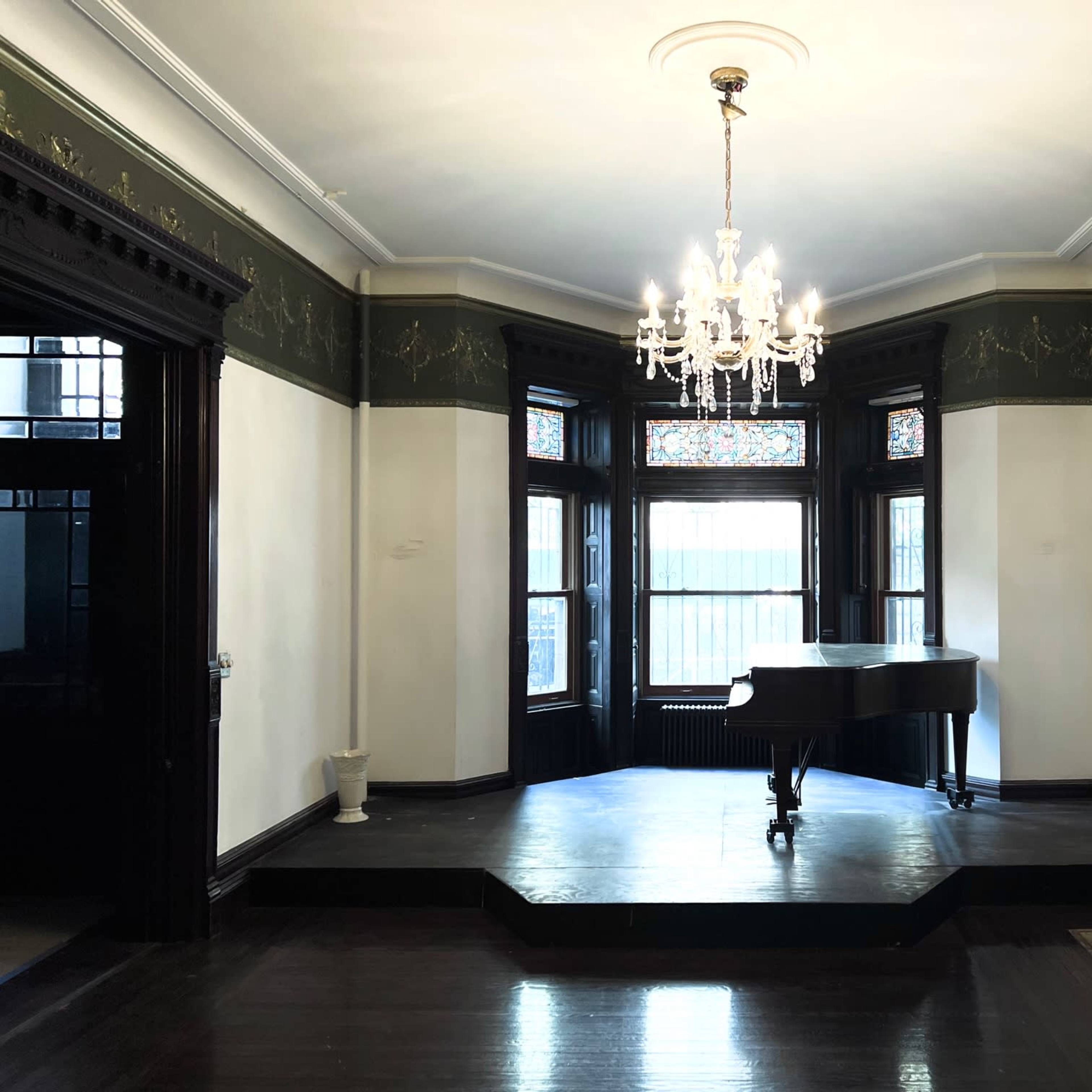 A grand room features a black piano on a raised platform, surrounded by large windows and ornate wall detailing.